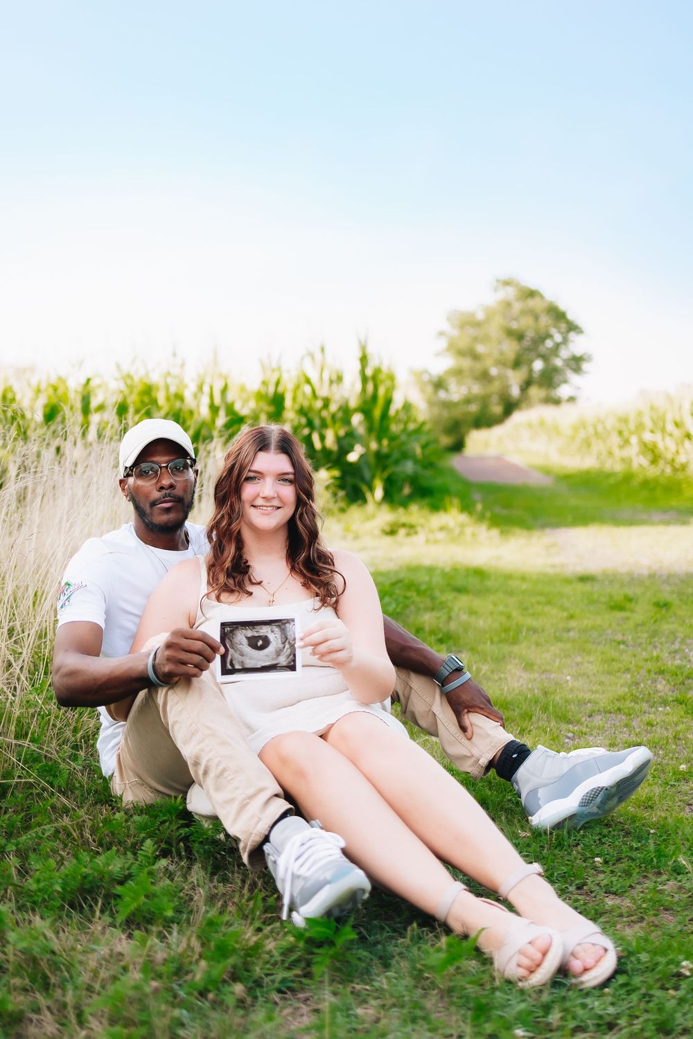 Couple sits in grassy field sharing ultrasound photo during pregnancy announcement shoot.