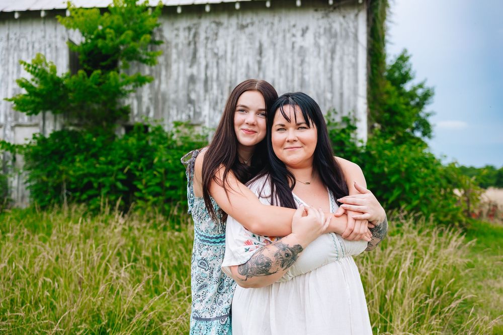 Two people embrace warmly against a scenic outdoor backdrop with lush green foliage.