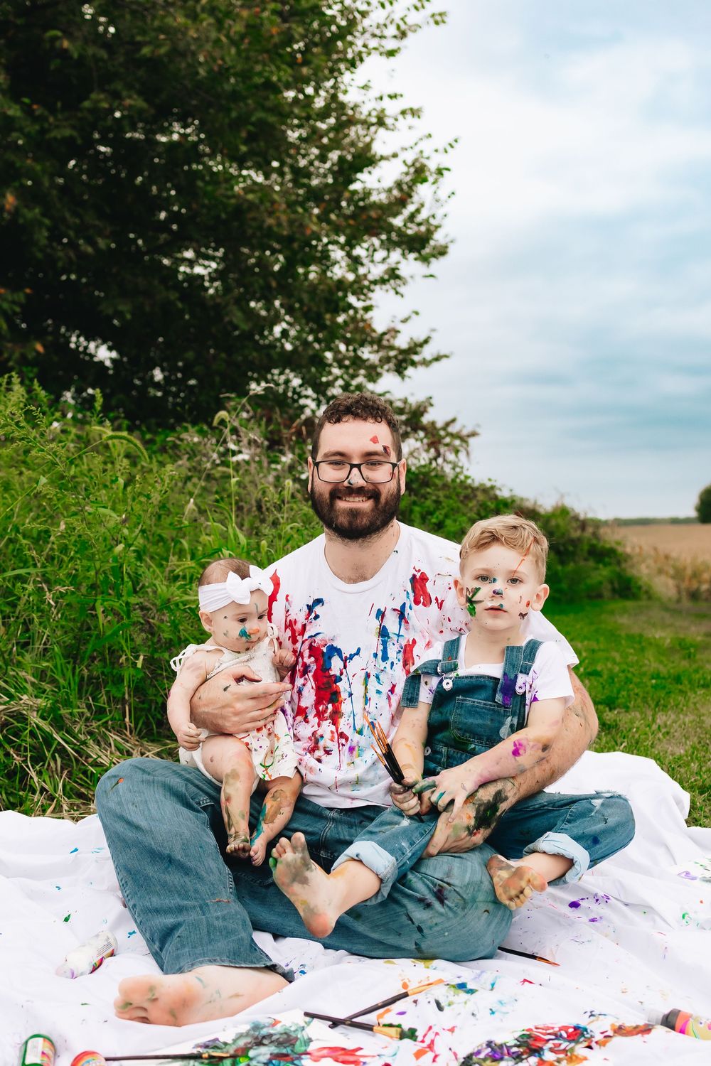 Parent and children covered in paint sit on blanket during outdoor art session.