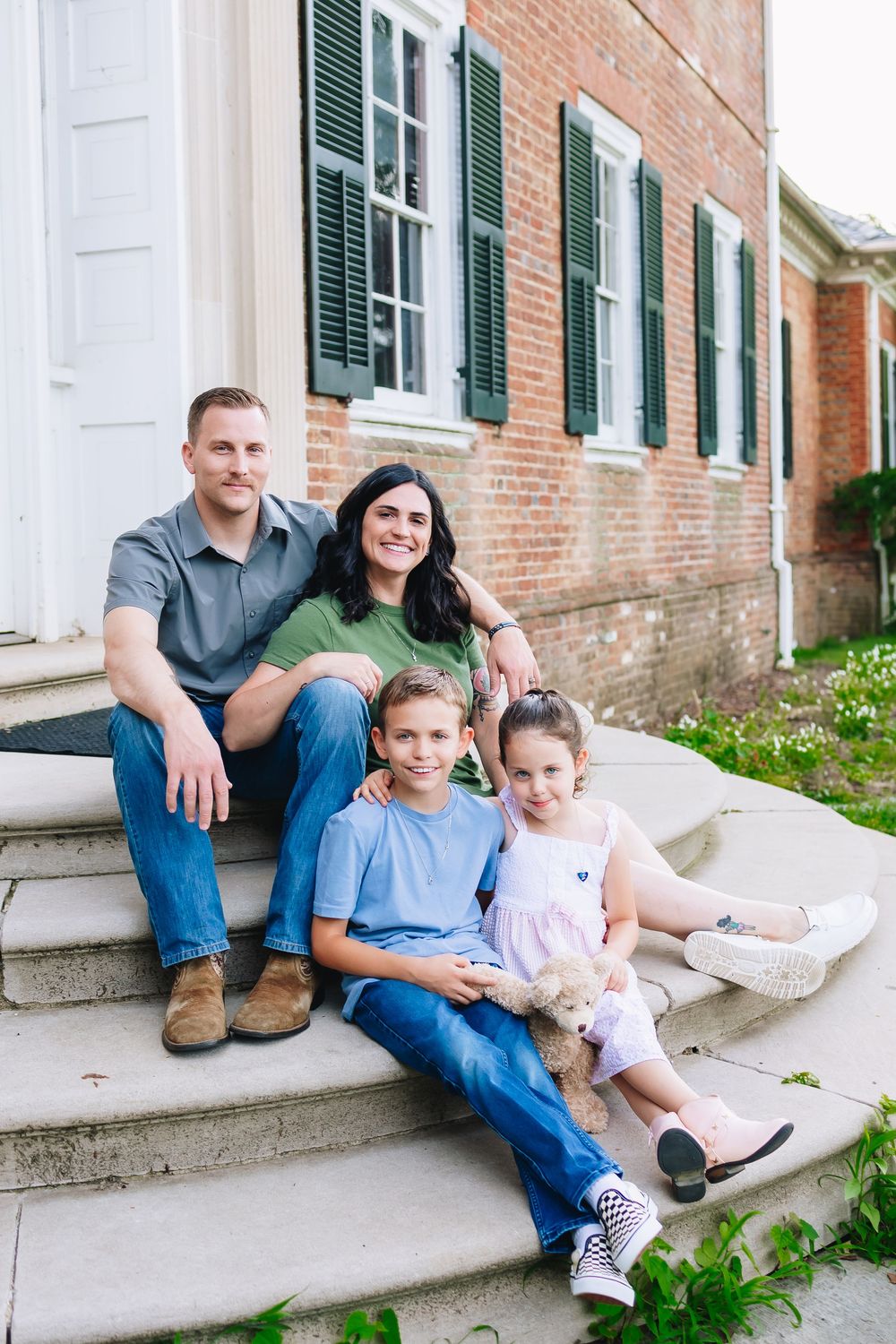 A family sits together on brick building steps surrounded by greenery on a sunny day.