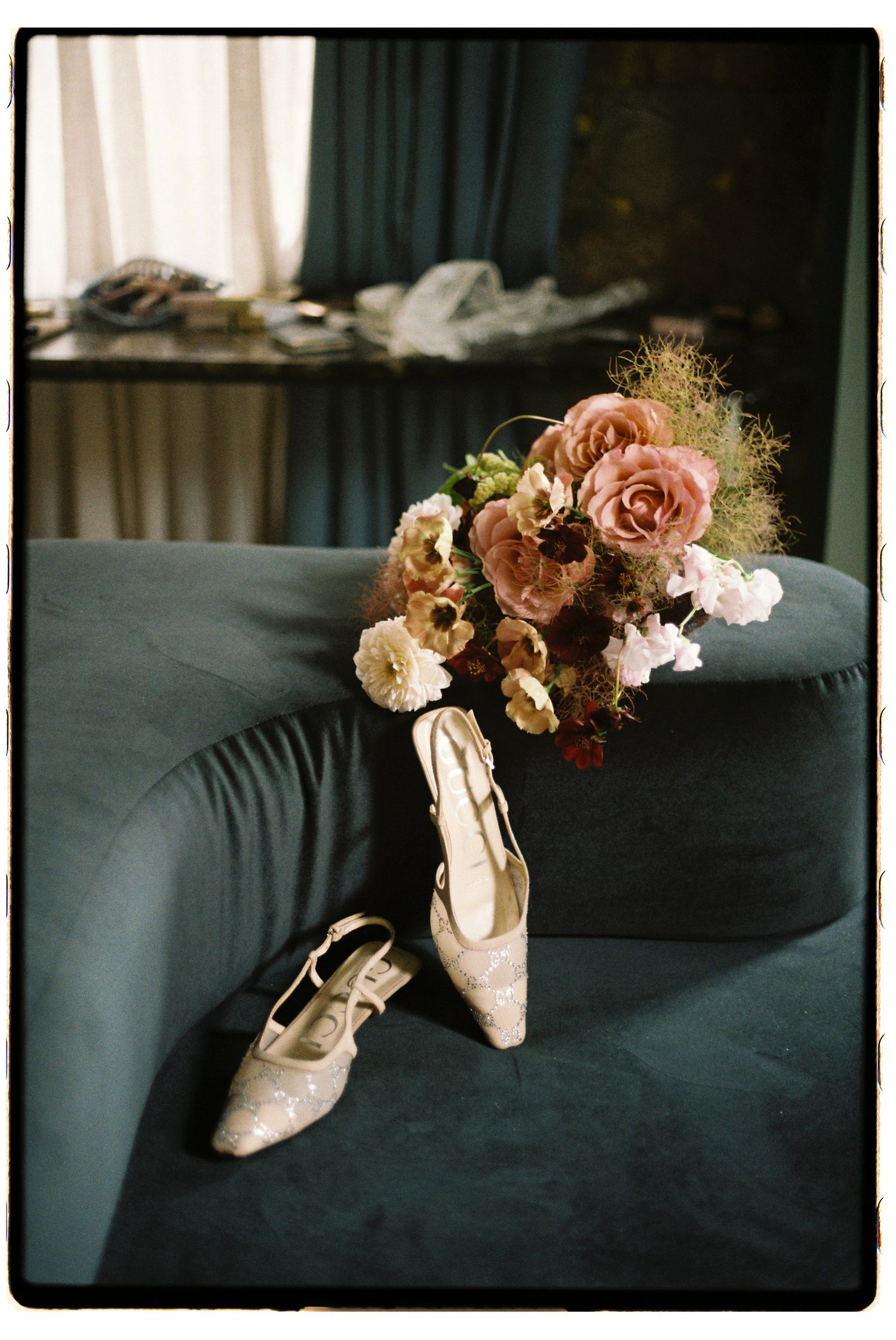 Pink ballet pointe shoes and pastel flowers arranged on a dark teal velvet couch in moody lighting.