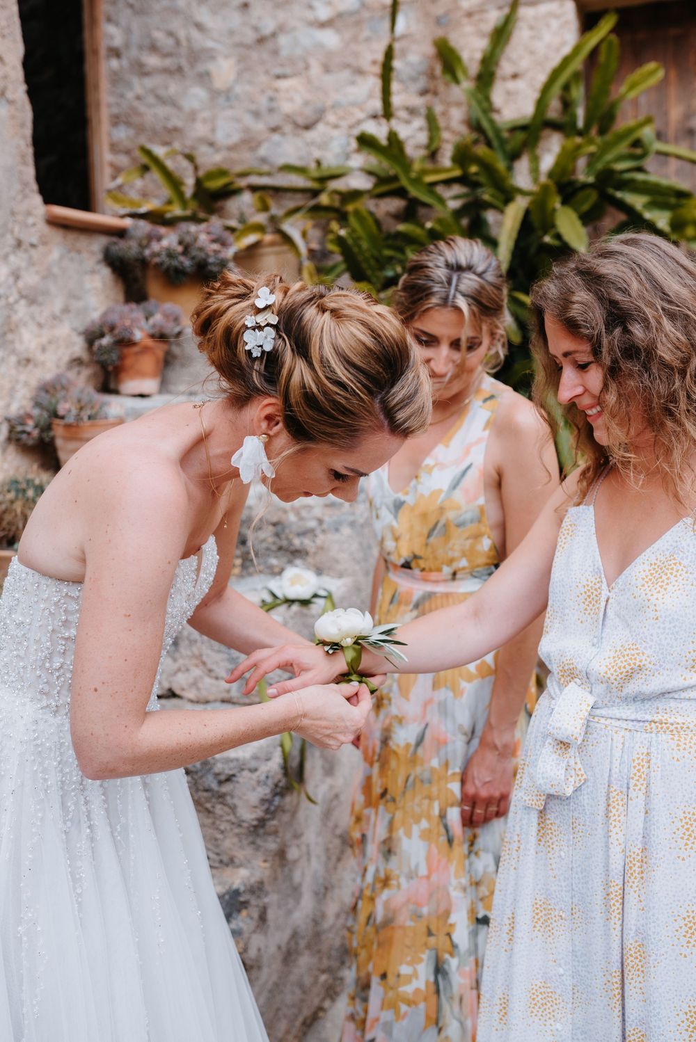 Die Braut bindet ihrer Brautjungfer das Blumenarmband um. Sie stehen im mallorquinischen Innenhof der Finca Xiclati auf Mallorca.