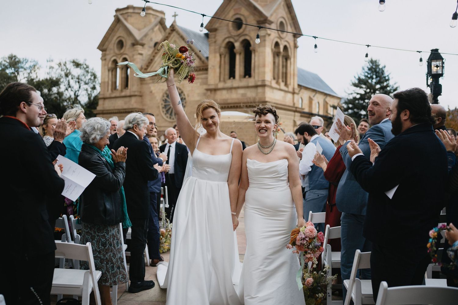 Two brides celebrate after outdoor wedding ceremony near historic stone cathedral with cheering guests. LGBT documentary photographer
