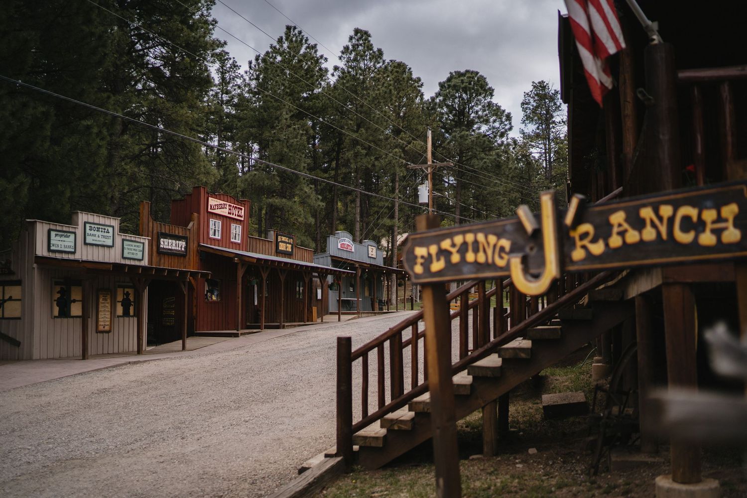 Rustic Western street at Flying J Ranch with wooden storefronts and pine trees under cloudy skies.