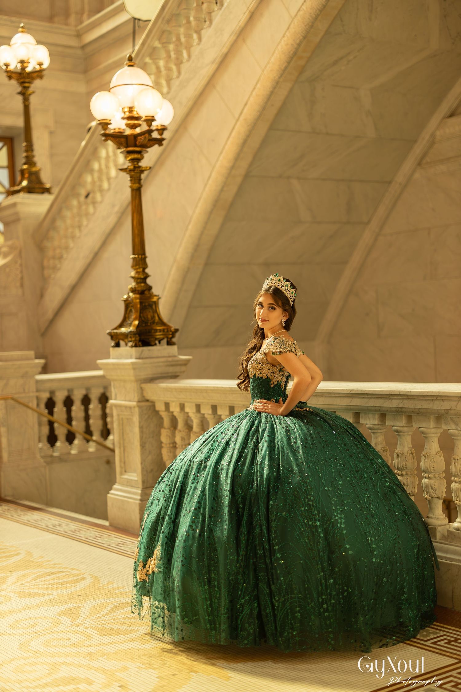 A person in a sparkling emerald green ball gown with a full tulle skirt poses elegantly on marble stairs.