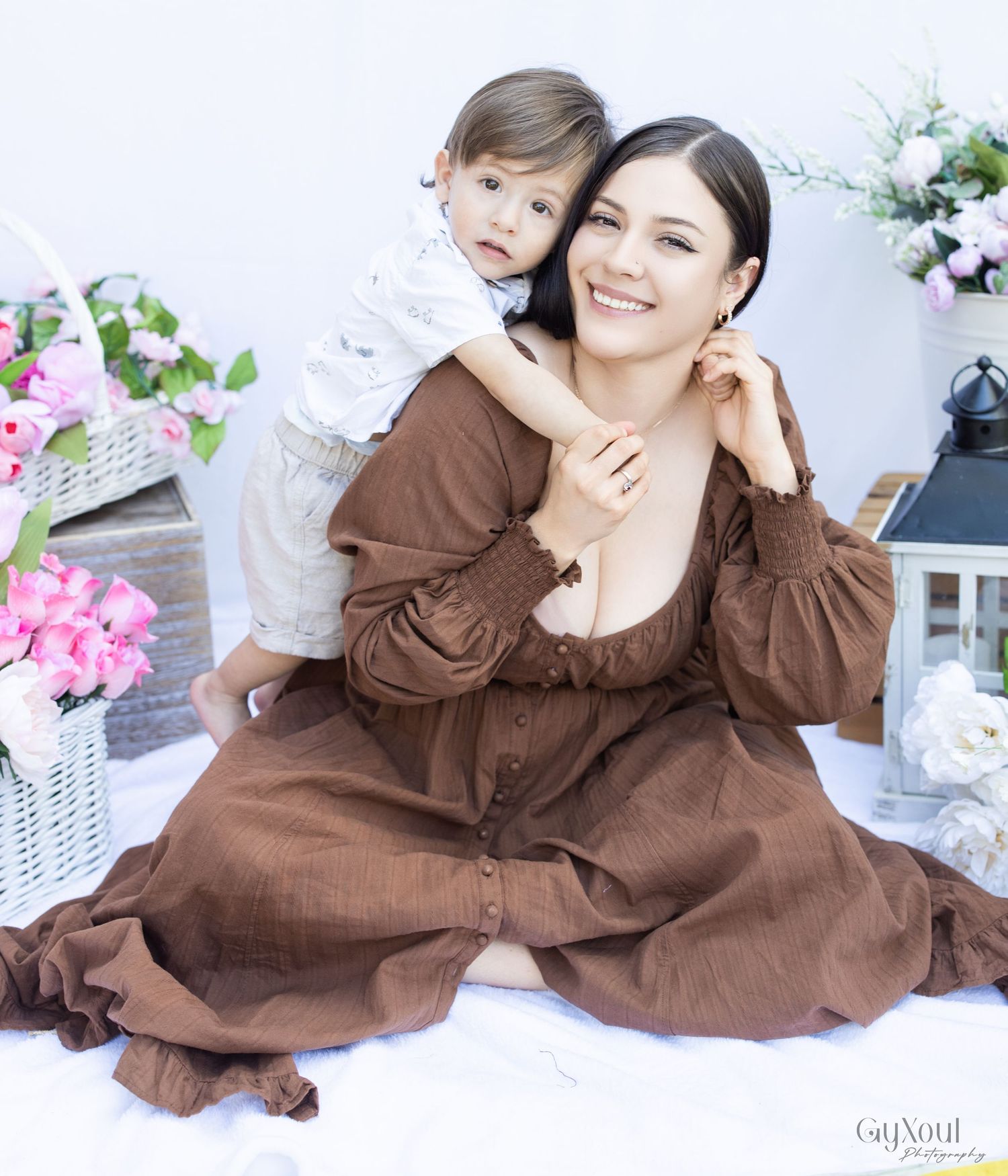 Two people in brown loungewear outfits pose casually on light-colored bedding with pink flowers in the background.
