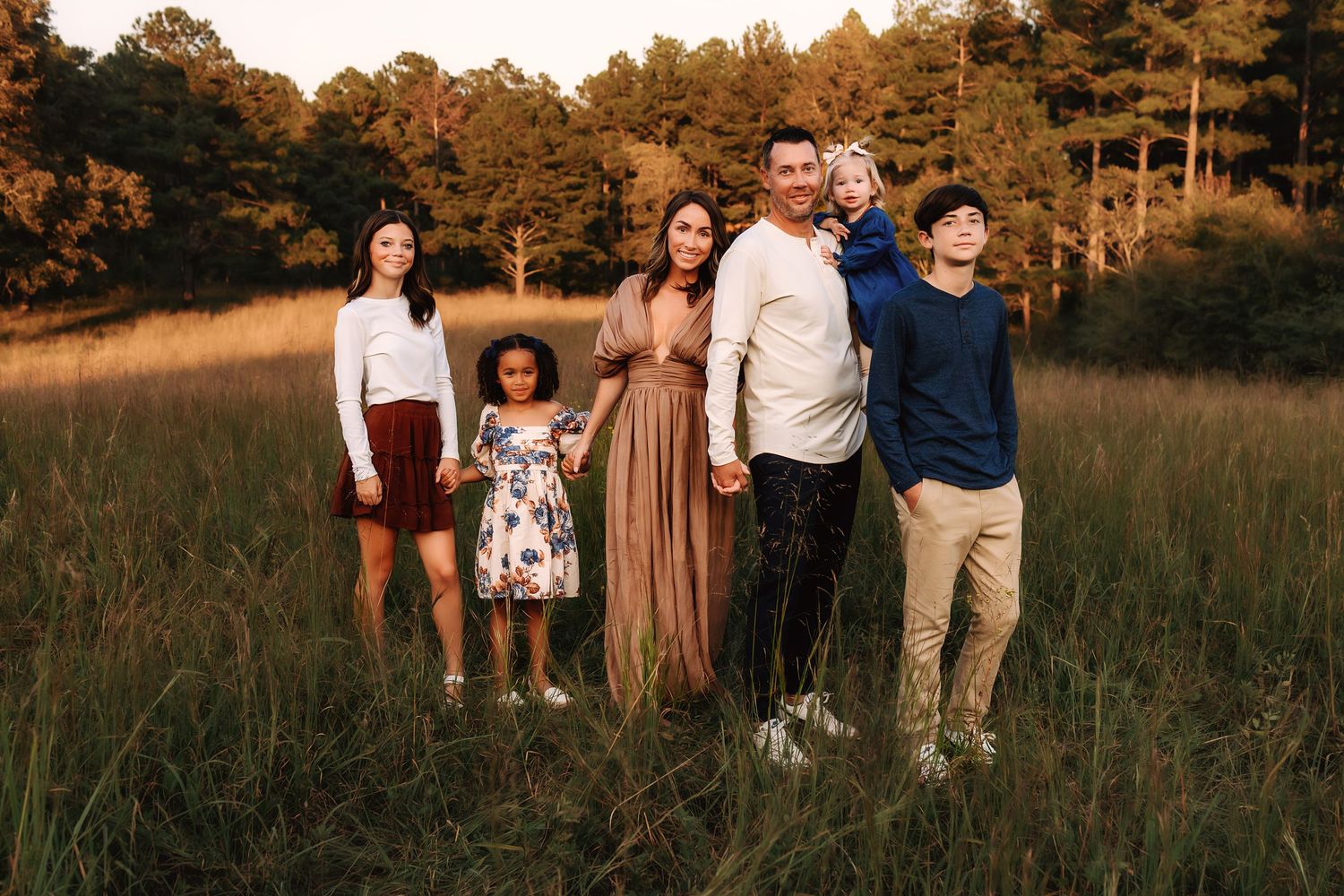 A group walking and posing together in a golden field at sunset with tall pine trees in the background.
