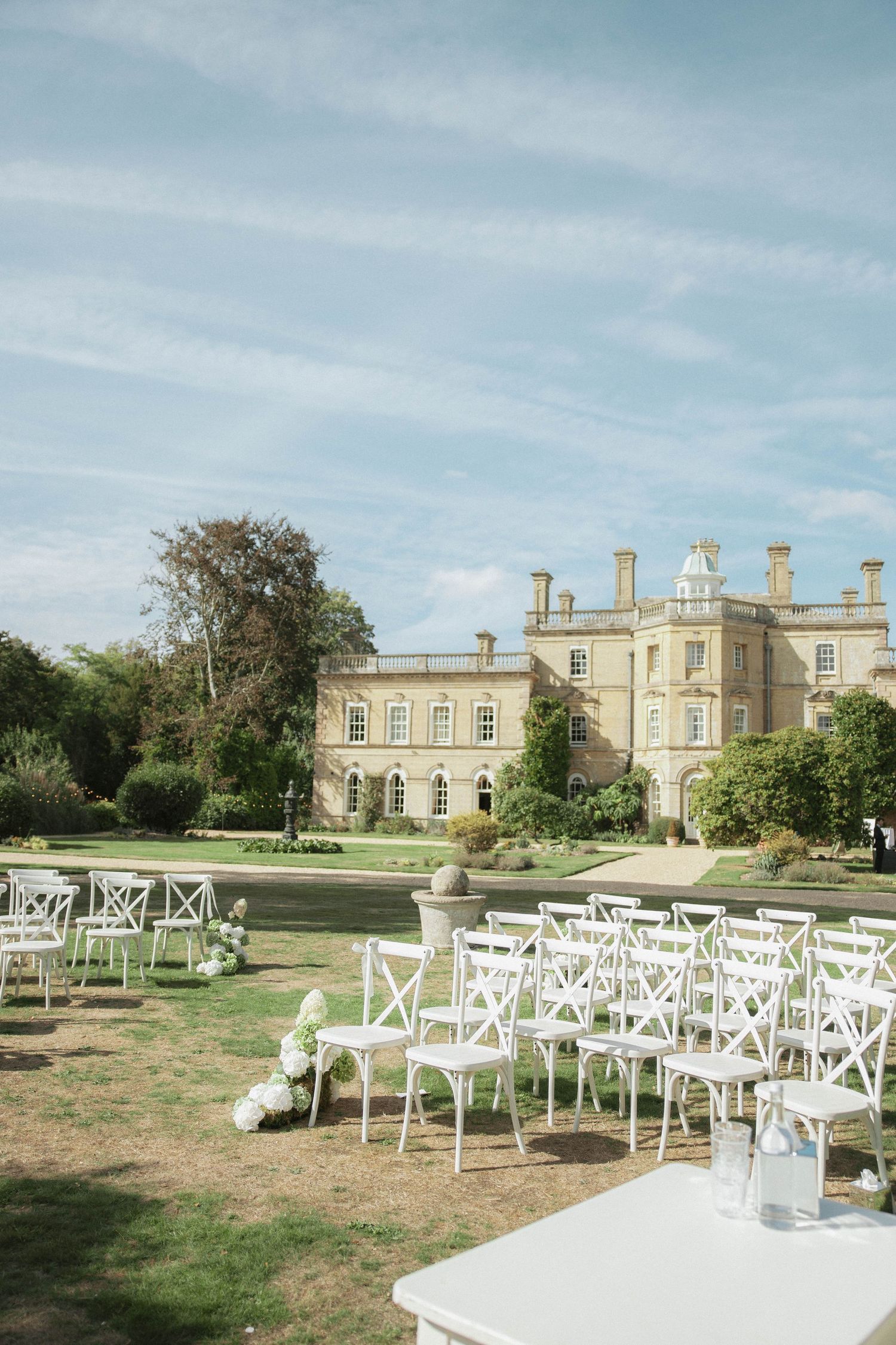 White cross-back chairs arranged in rows on a lawn before a grand English country house wedding ceremony on a sunny day.
