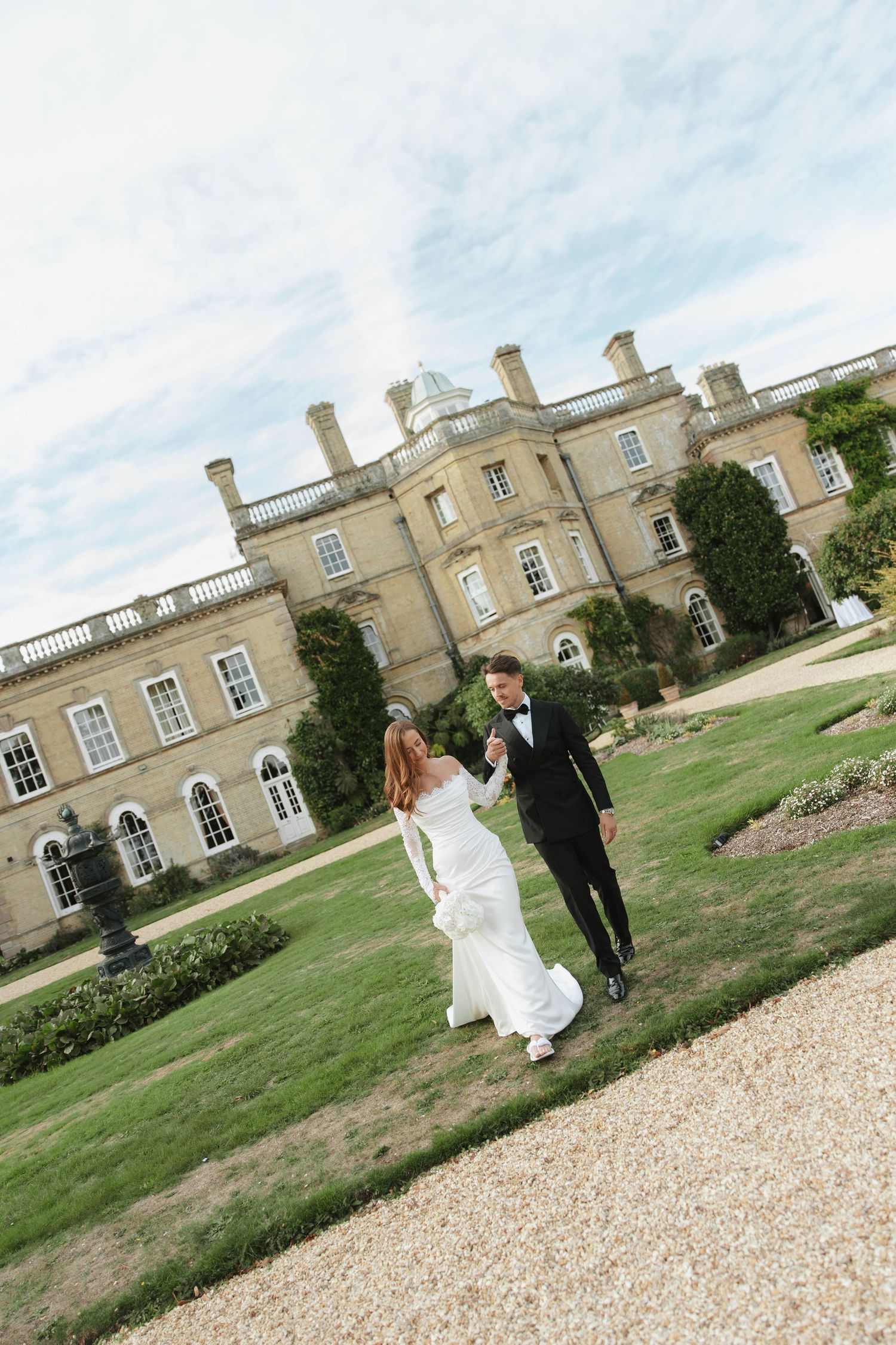 A bride and groom walk hand in hand across manicured gardens outside an elegant English country manor on their wedding day.