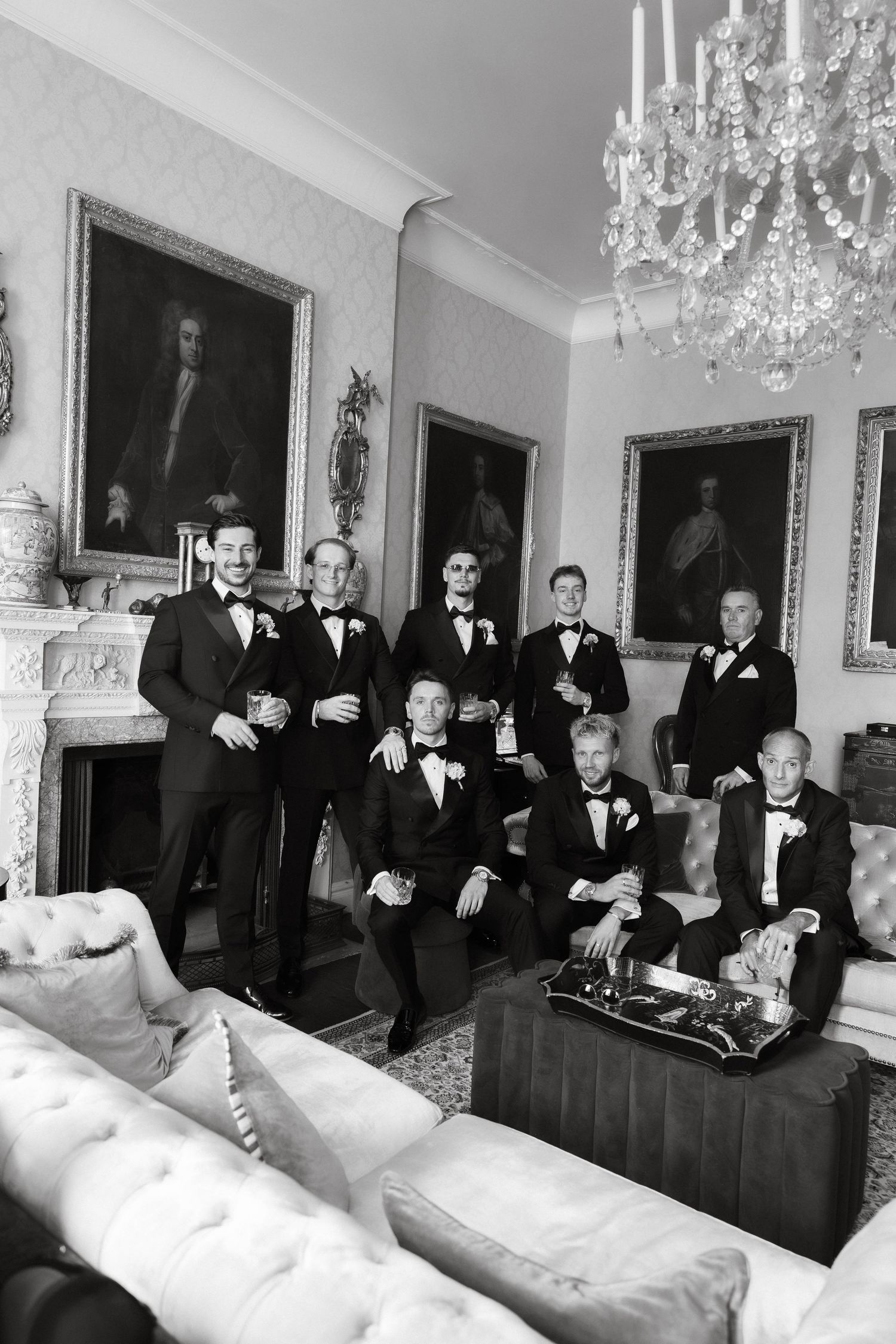 Black and white photo of groomsmen in tuxedos posing in an elegant room with chandelier and portrait paintings.