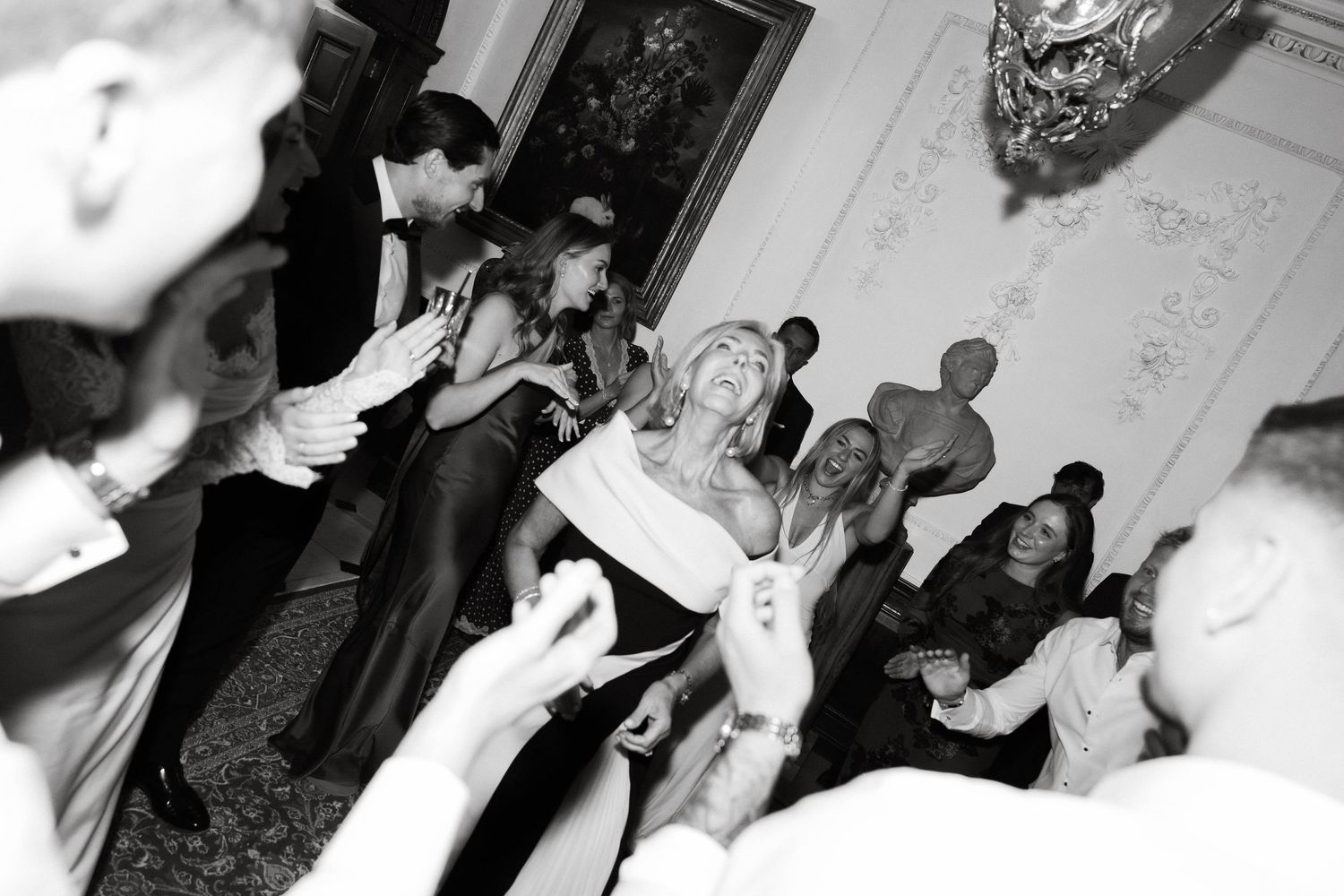 Black and white wedding reception photo of bride dancing joyfully surrounded by clapping guests in elegant attire.