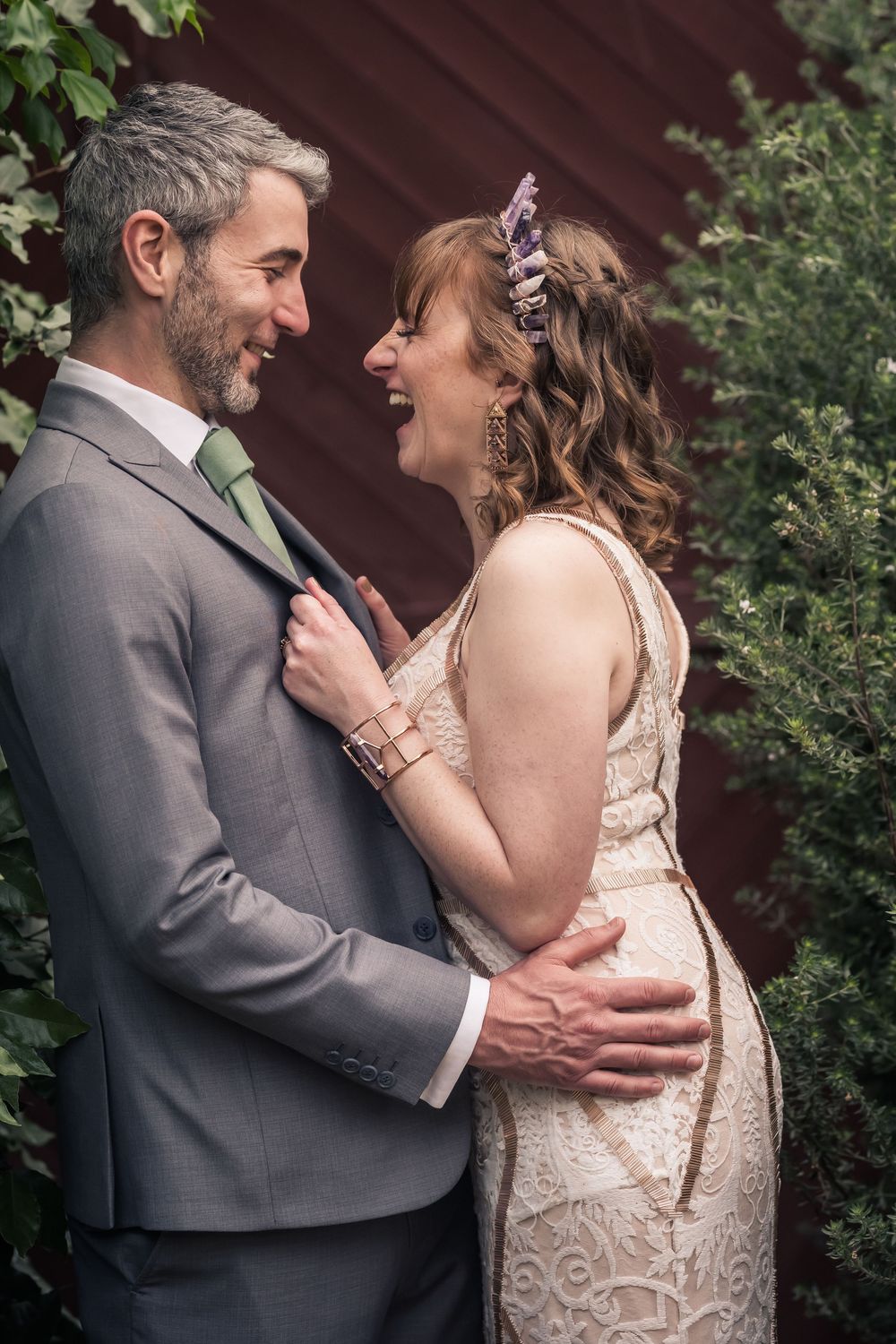 A couple shares a tender moment during an outdoor wedding photoshoot beside greenery and a rustic red wall.