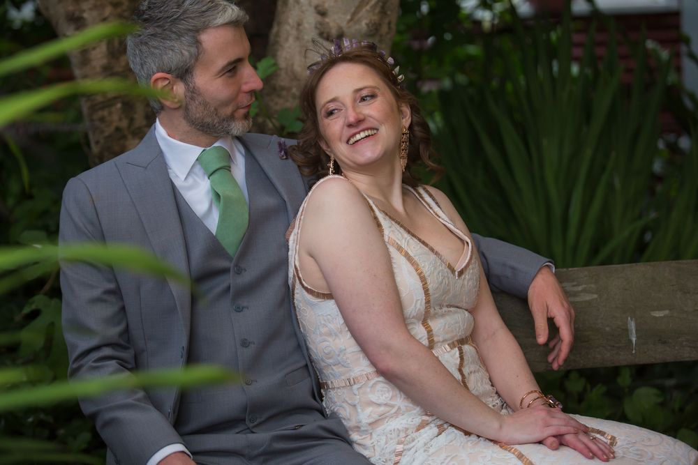 A couple in wedding attire sits together on a bench surrounded by tropical plants in an outdoor garden setting.