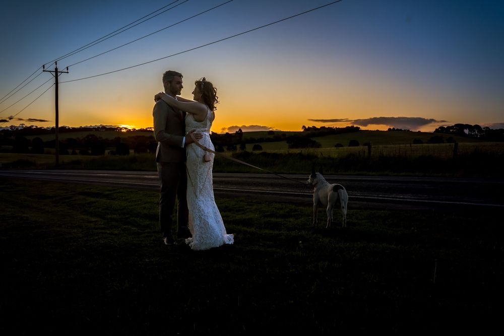 Silhouetted couple embraces at sunset in rural field with power lines and dog in background.