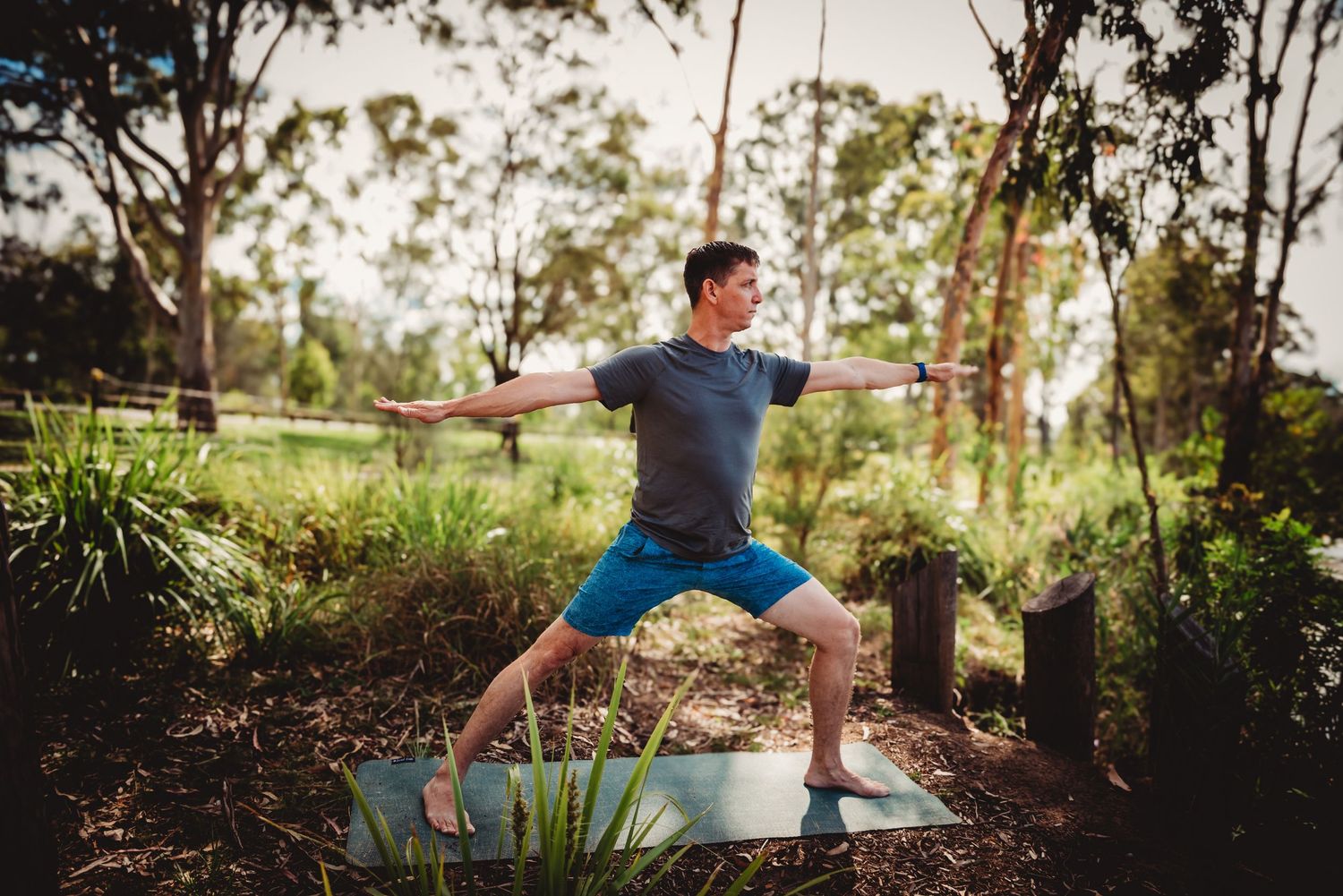 Someone practicing yoga in warrior pose on a mat outdoors in a lush garden setting during sunset.