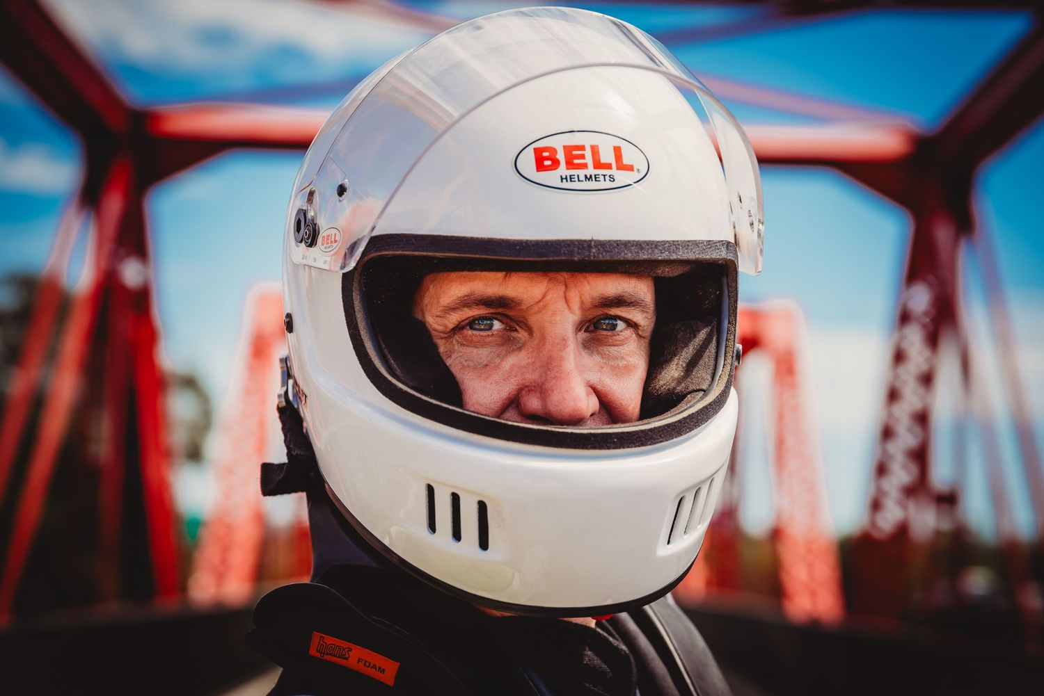 A motorcyclist wearing a white Bell helmet photographed against a red bridge structure in the background.