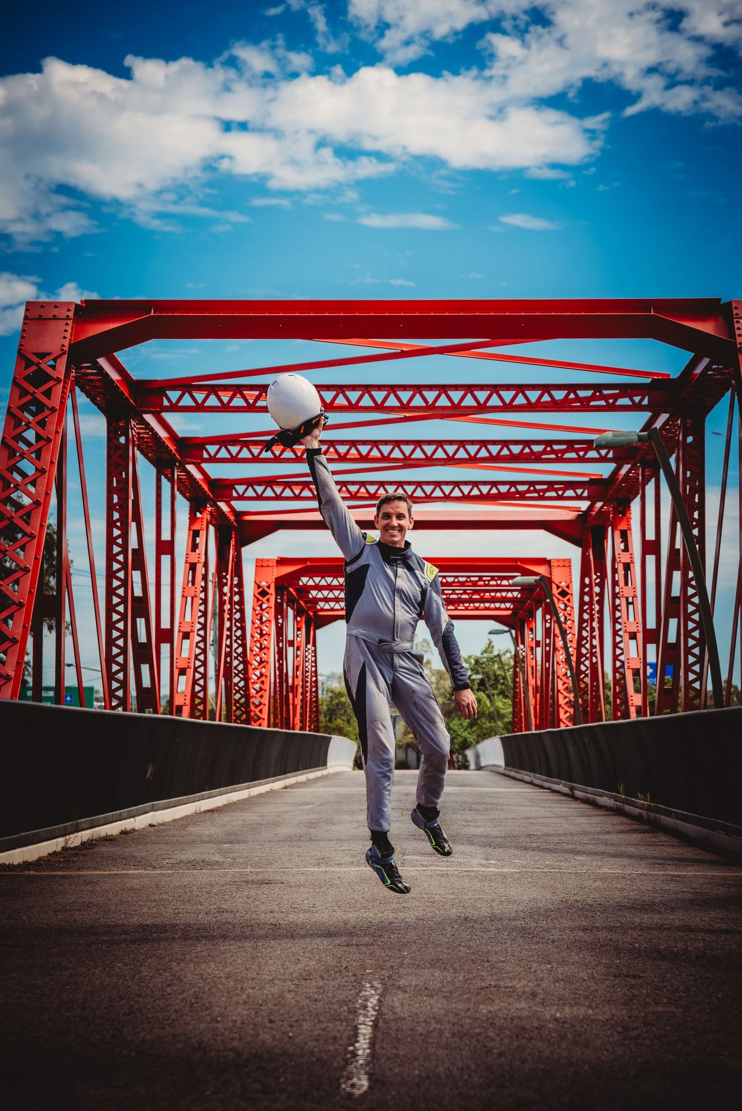Joyful person in grey suit jumping on red steel truss bridge against vibrant blue sky with white clouds.