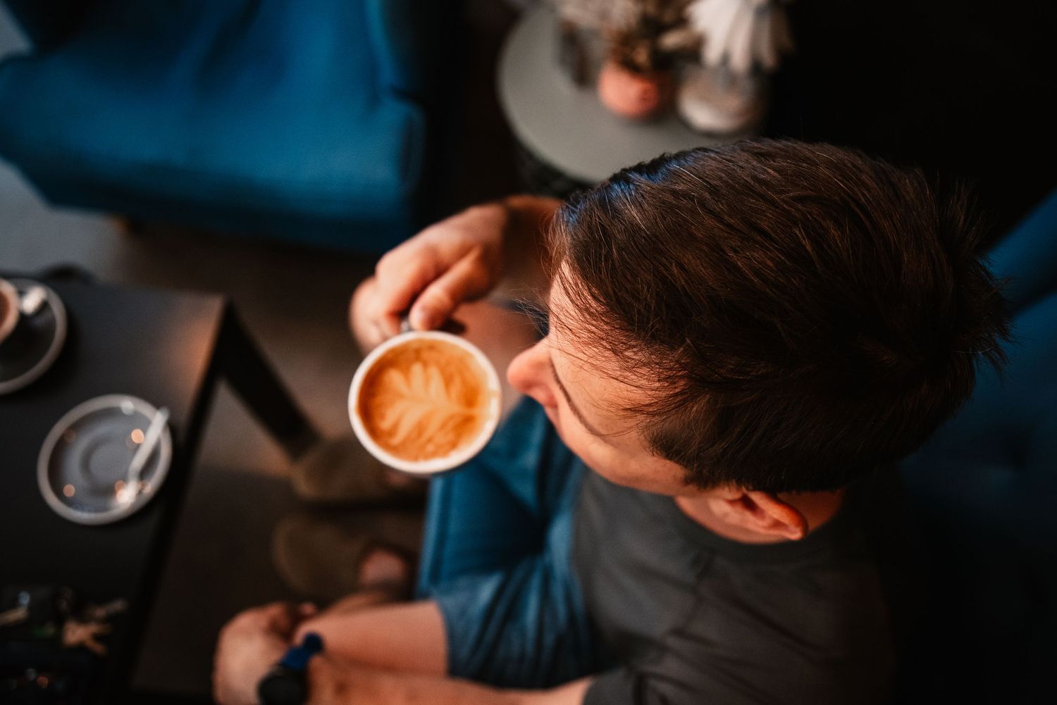 Overhead view of someone in a dark shirt drinking a latte with artistic foam design in a cafe.