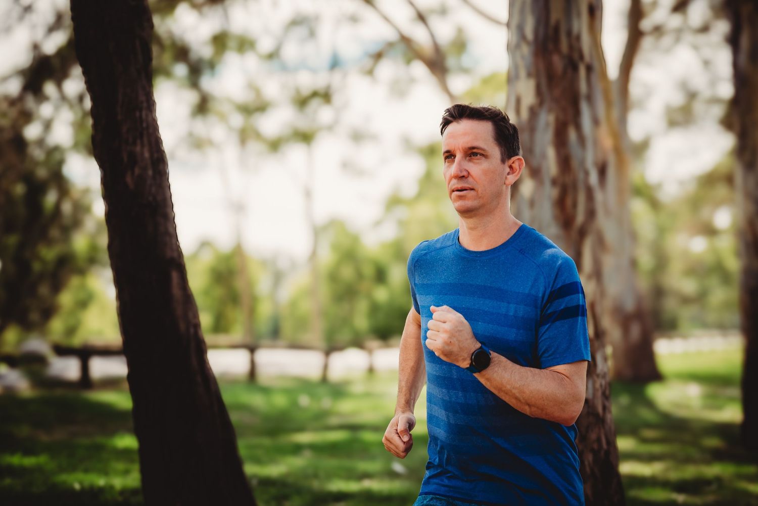 Person in blue shirt running and jogging through scenic wooded park with trees during outdoor workout.