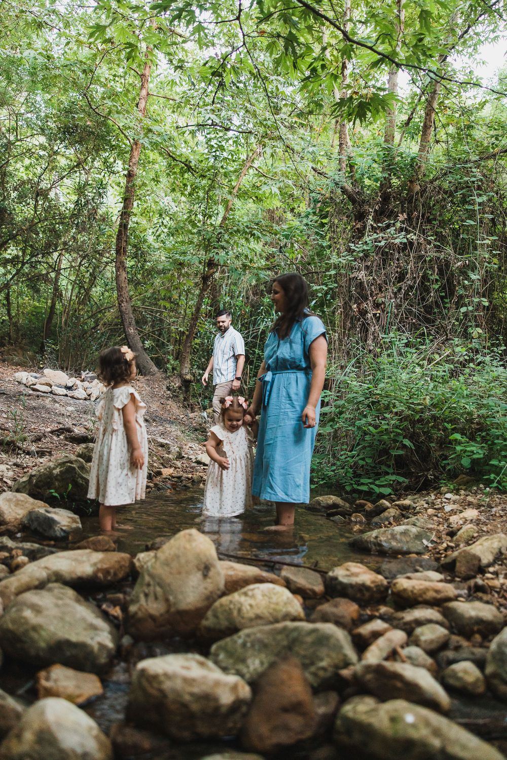 People stand by a rocky stream in a lush forest setting wearing dresses and light summer clothing.