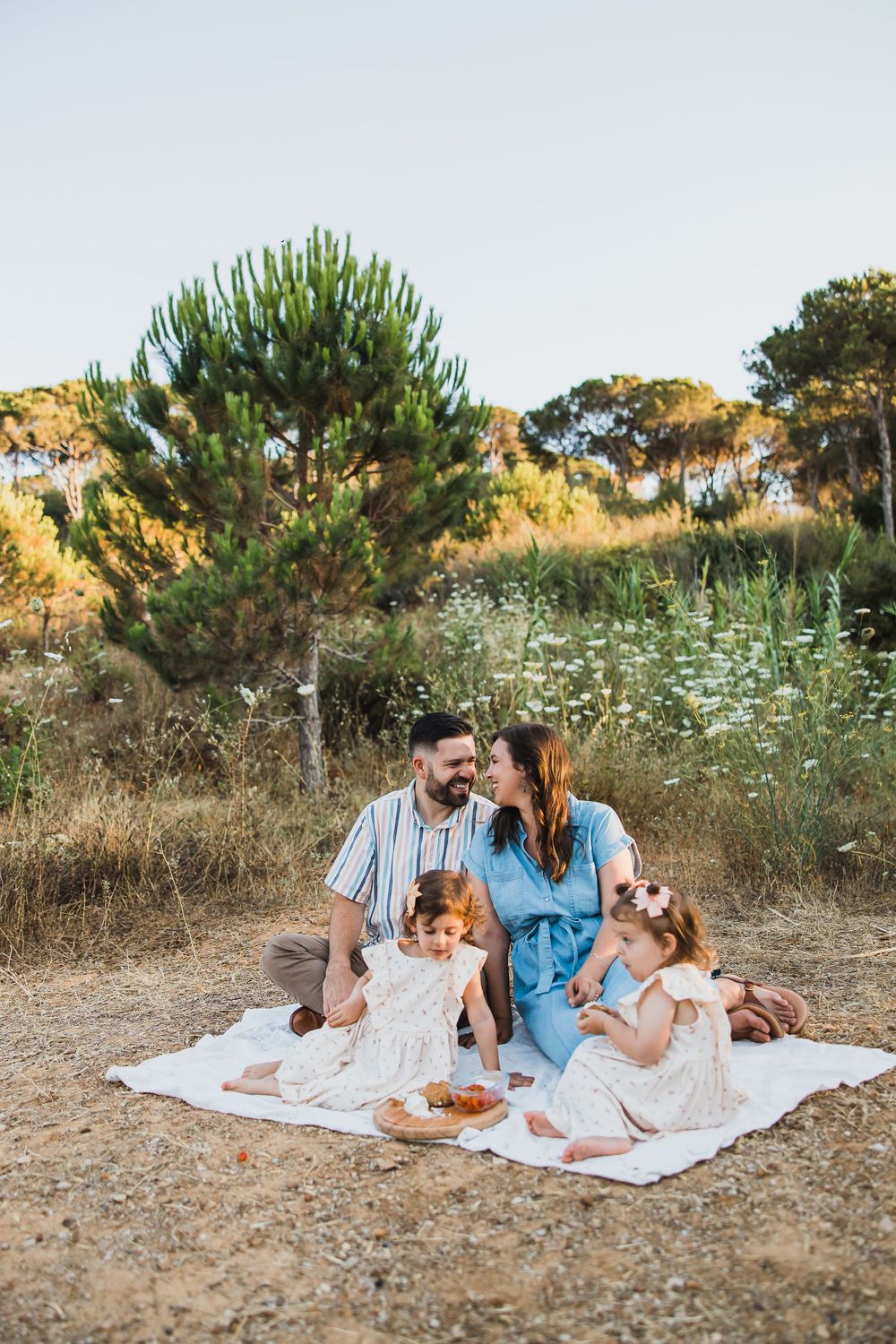 Family enjoying a picnic on a blanket outdoors in a naturalistic setting surrounded by pine trees and wildflowers.
