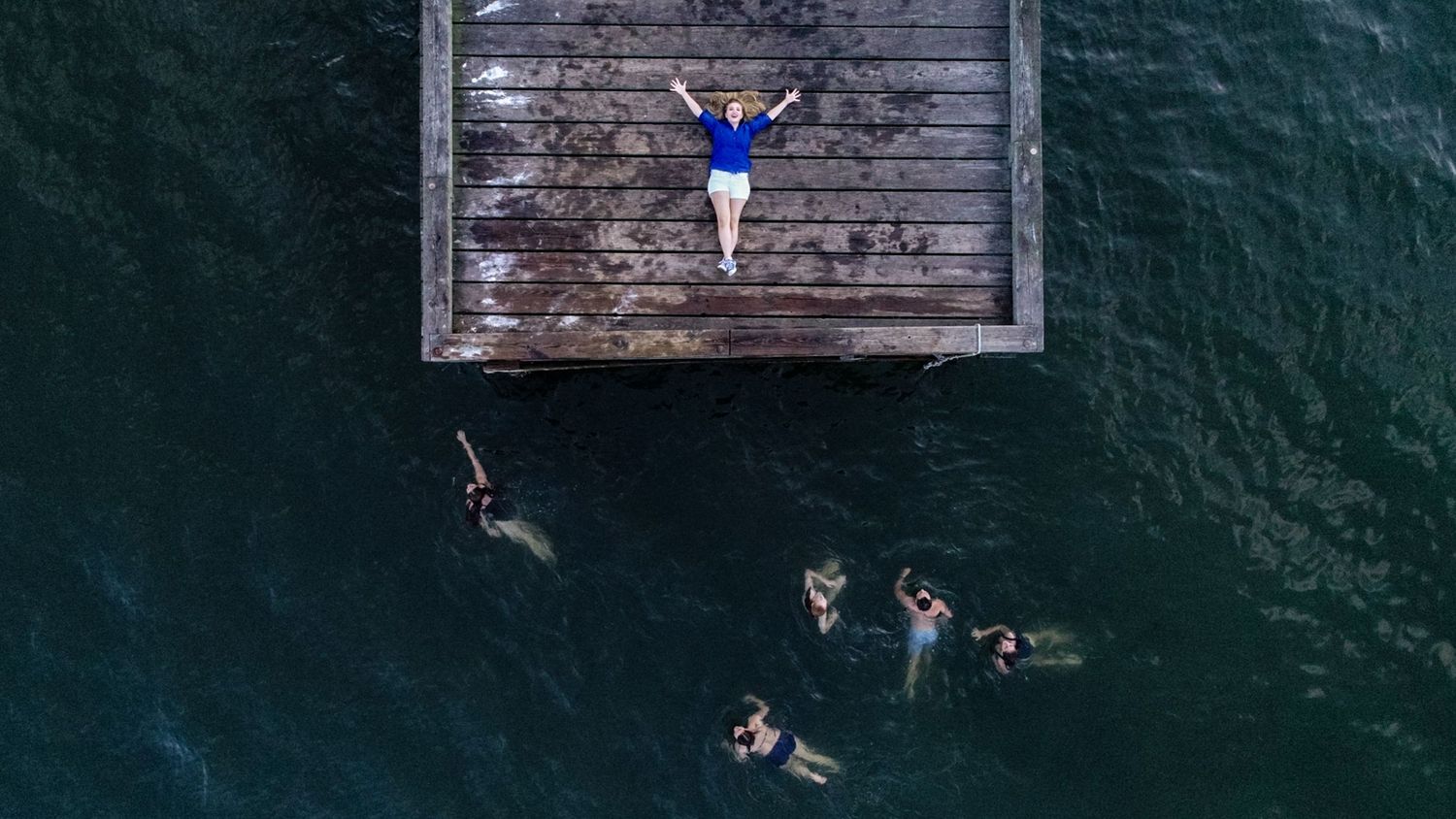 Aerial view of people jumping from a long wooden pier into dark blue ocean waters on a sunny day.