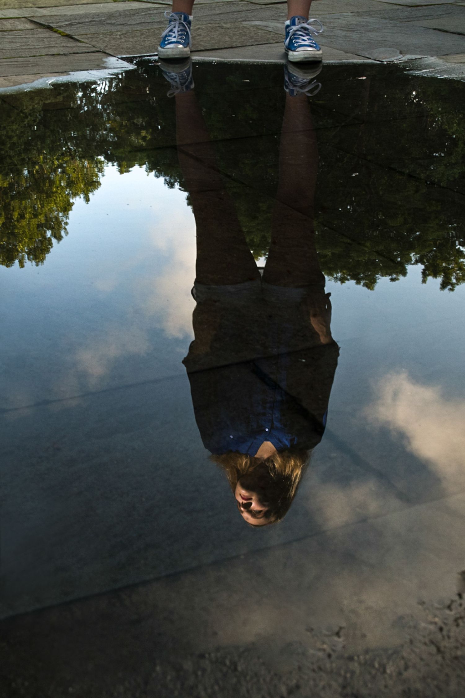 Upside down reflection of a person in a puddle showing dark water and cloudy sky background.