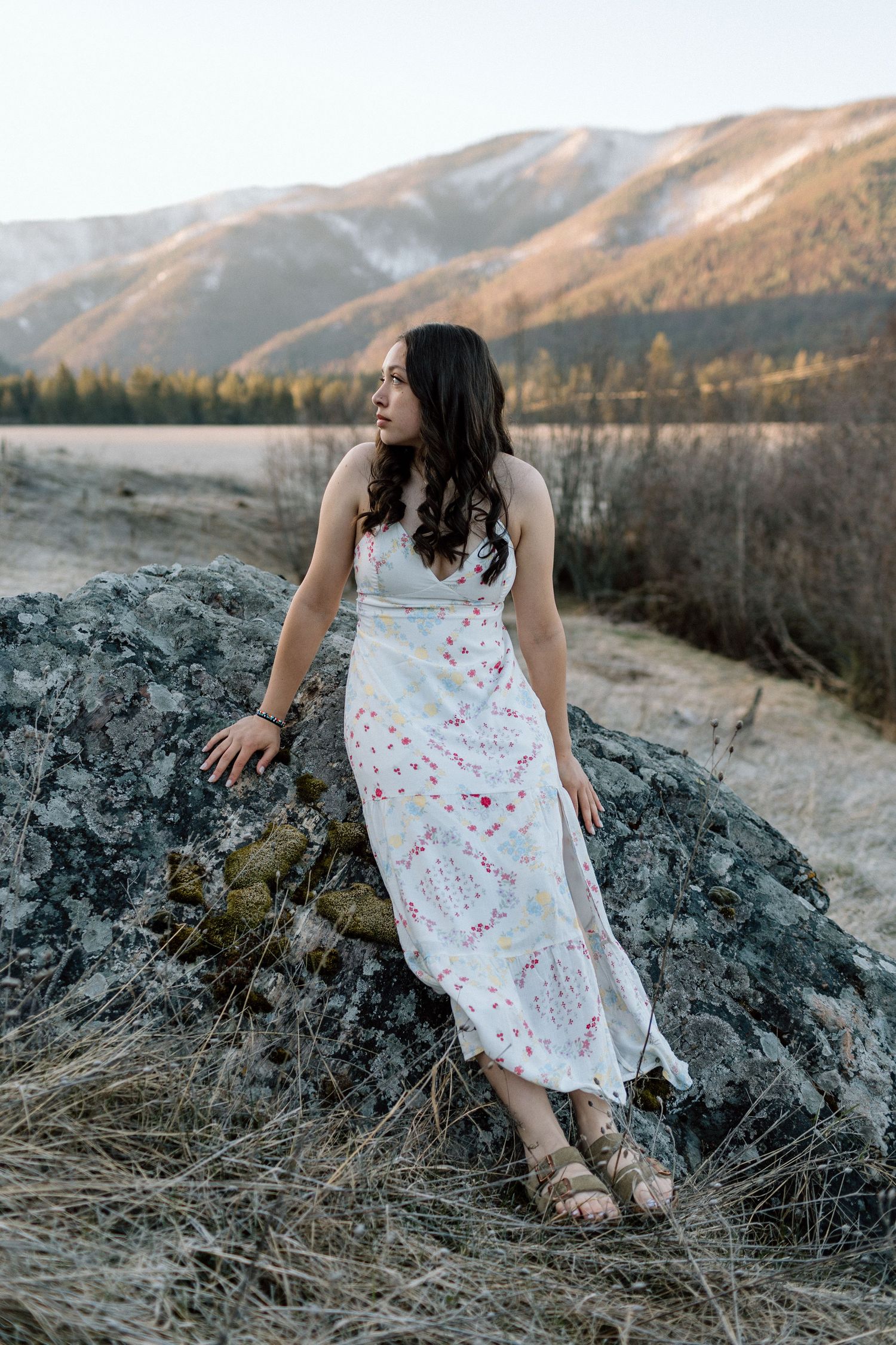 A floral white dress stands out against mountain scenery as the subject poses on dark rocks at sunset.