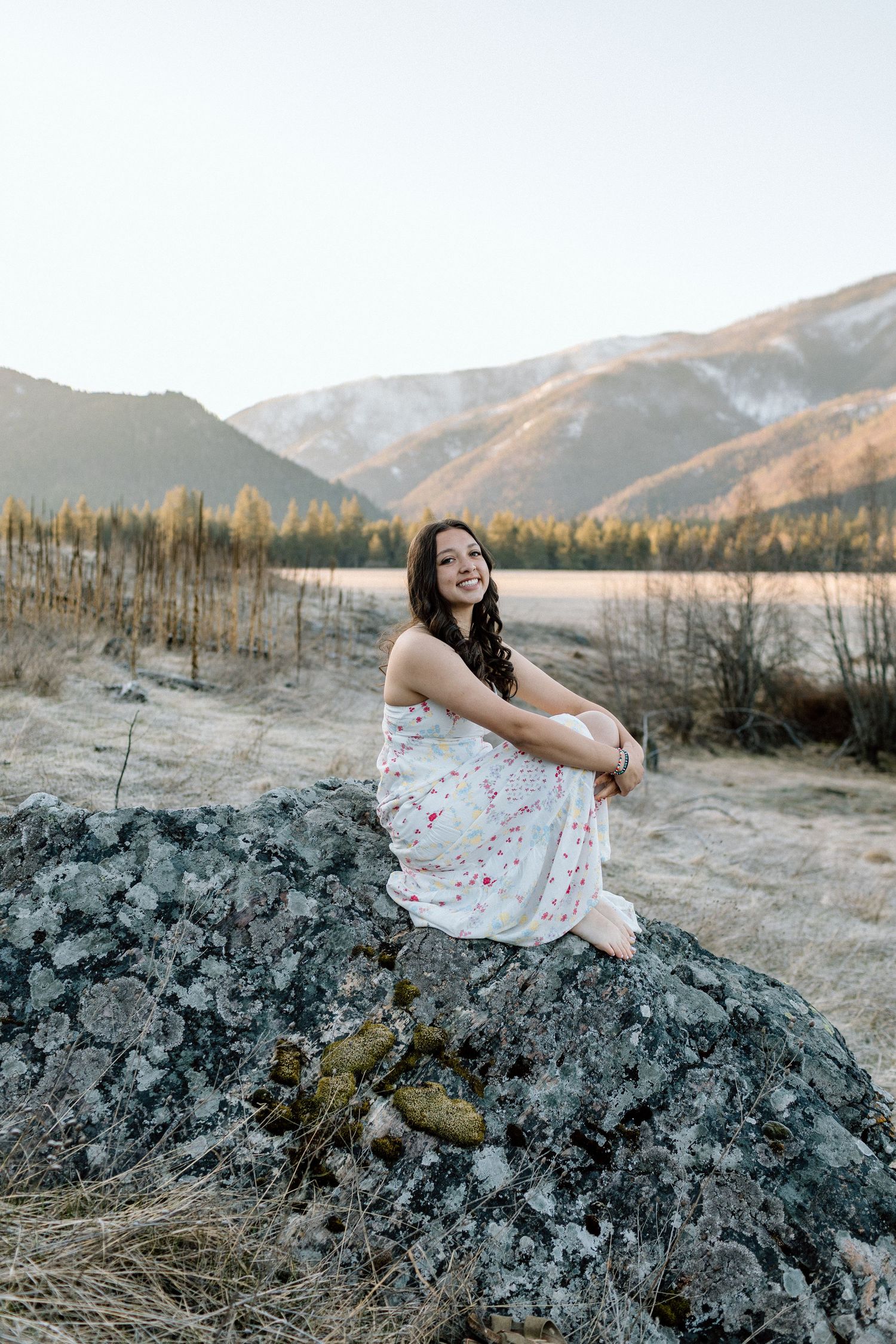 White floral dress contrasts with dark rocks as mountains and trees create a scenic backdrop.