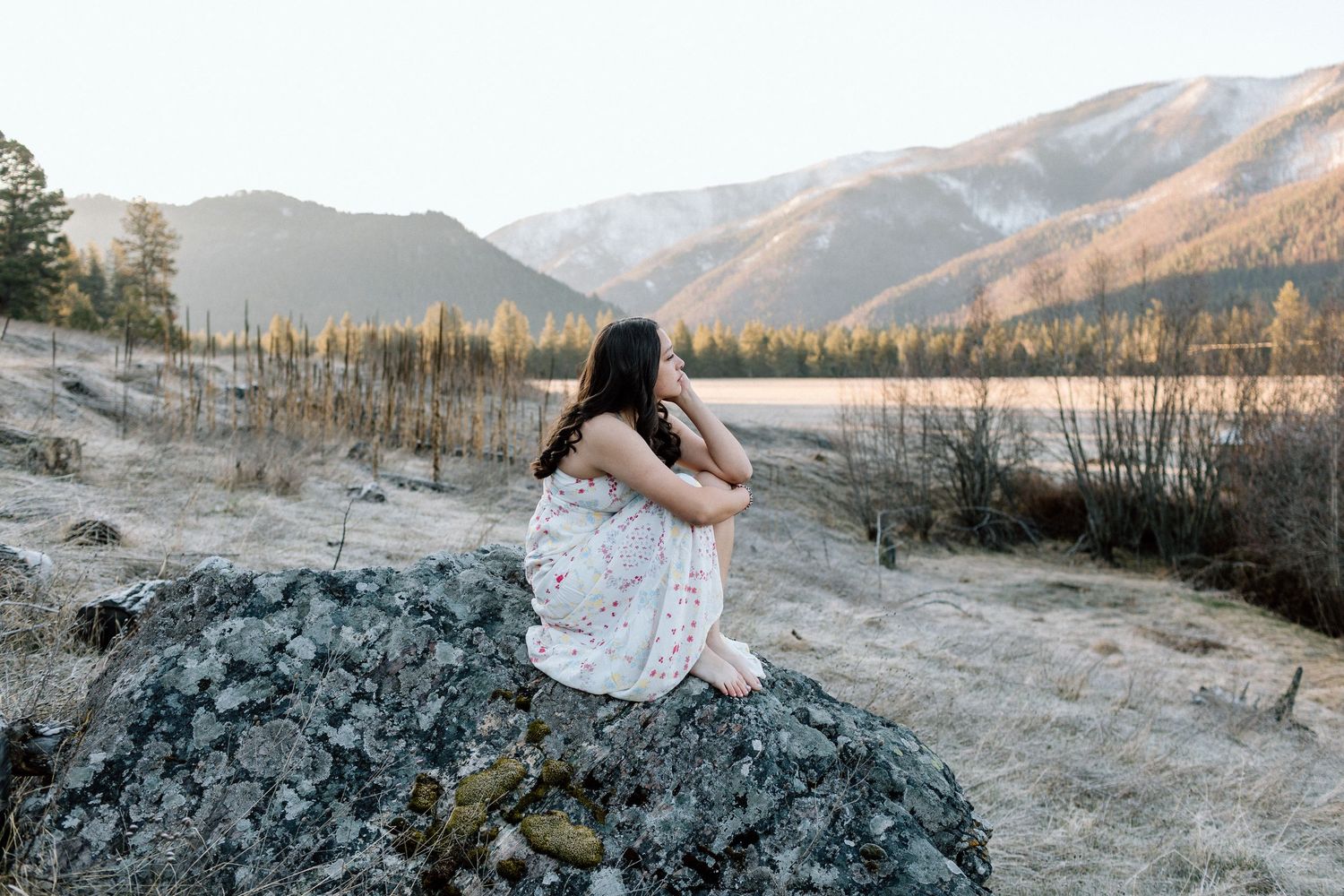 Figure in white dress sits contemplatively on rocky outcrop with mountain vista behind.