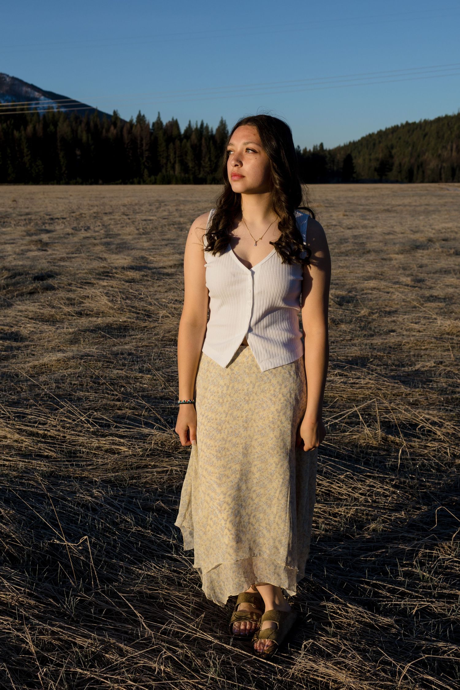 Person in light colored outfit stands in dry field with mountains and forest in background.
