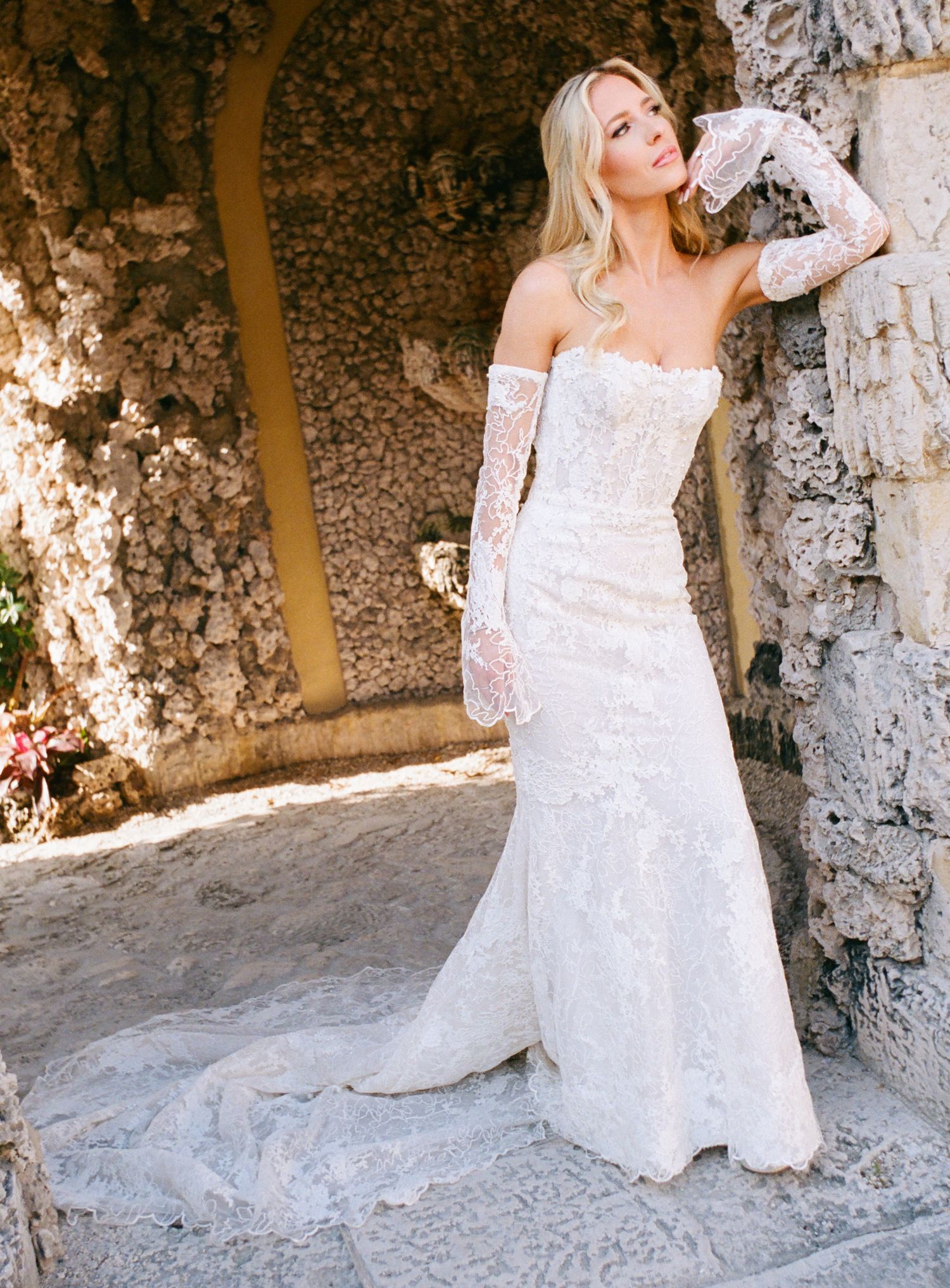 Elegant lace wedding gown with off-shoulder sleeves and fitted silhouette photographed against stone archway in natural light.