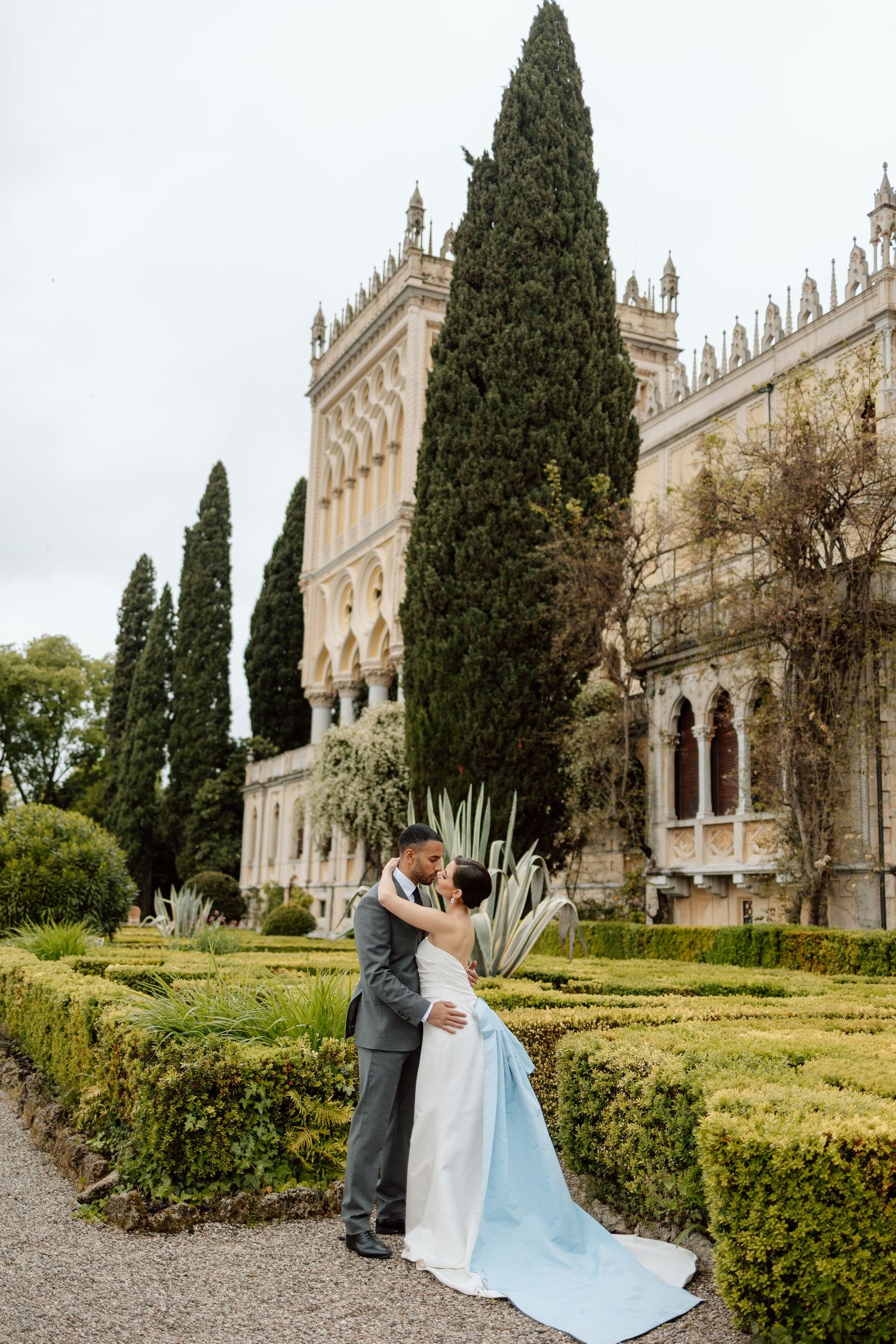 A couple embraces in a garden at a grand European estate with Italian cypress trees and architecture. Destination wedding photographer.
