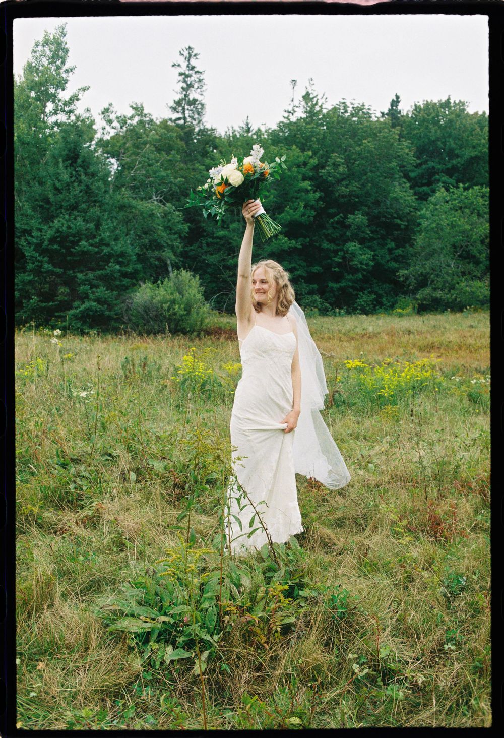 Bride stands in field holding up bouquet in celebration during bridal party photos at a coastal wedding in Downeast Maine.