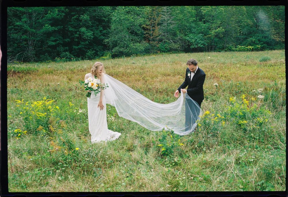 Young couple stands in a green field while he holds her veil and she looks back at him for this coastal summer wedding in downeast Maine.