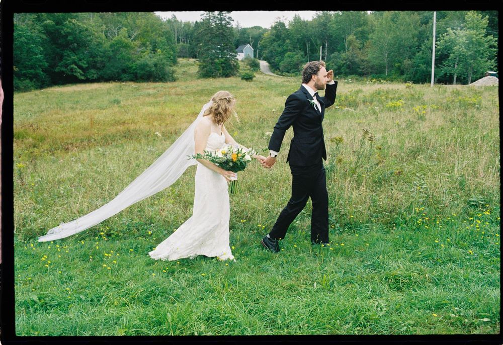 Couple walking through field and playfully holding hands and looking forward on color film in a downeast wedding in Brooklyn, Maine.