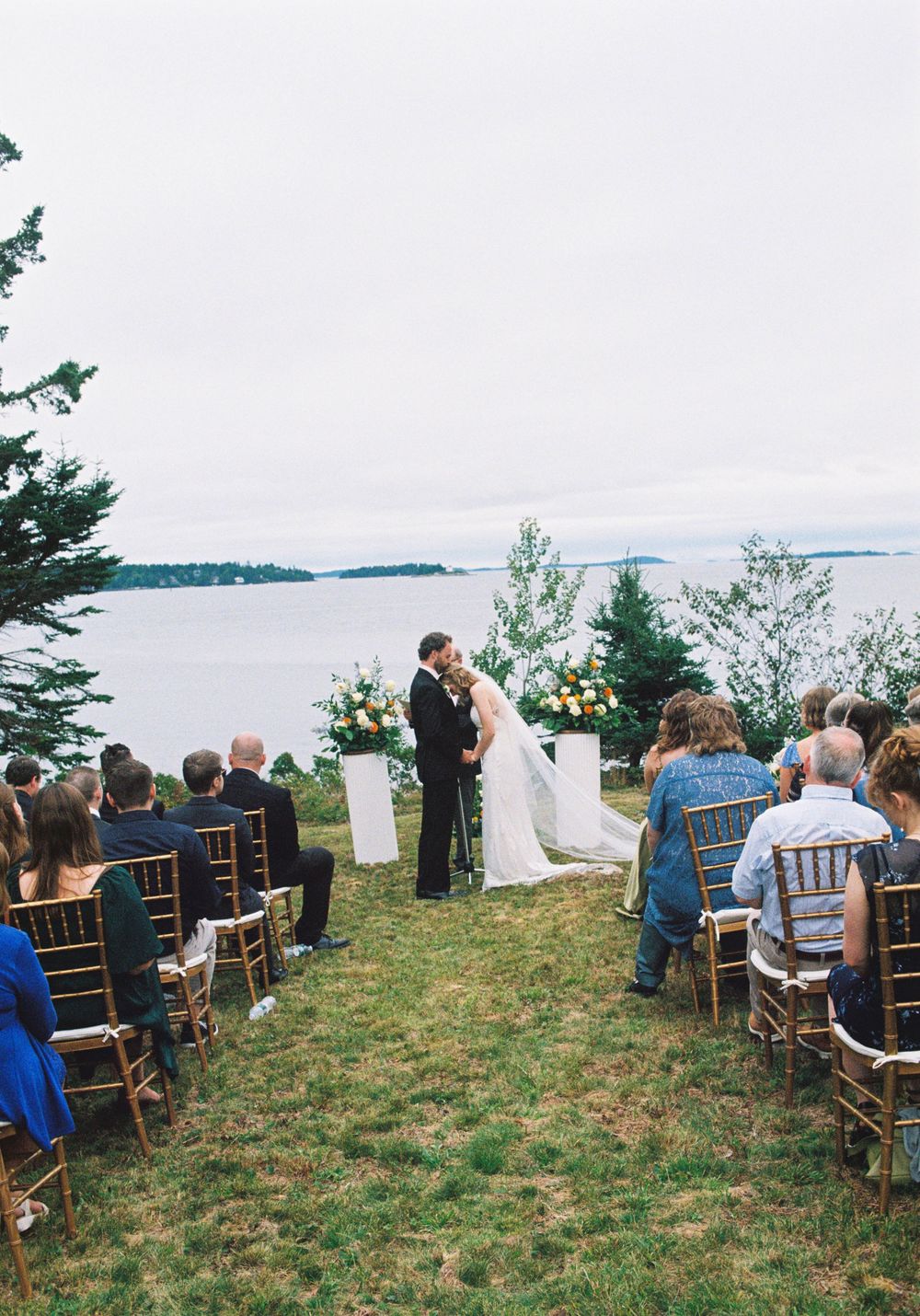 Photo of the wedding ceremony of a young couple as all their guests look on, the groom kisses the bride on the forehead by the sea.