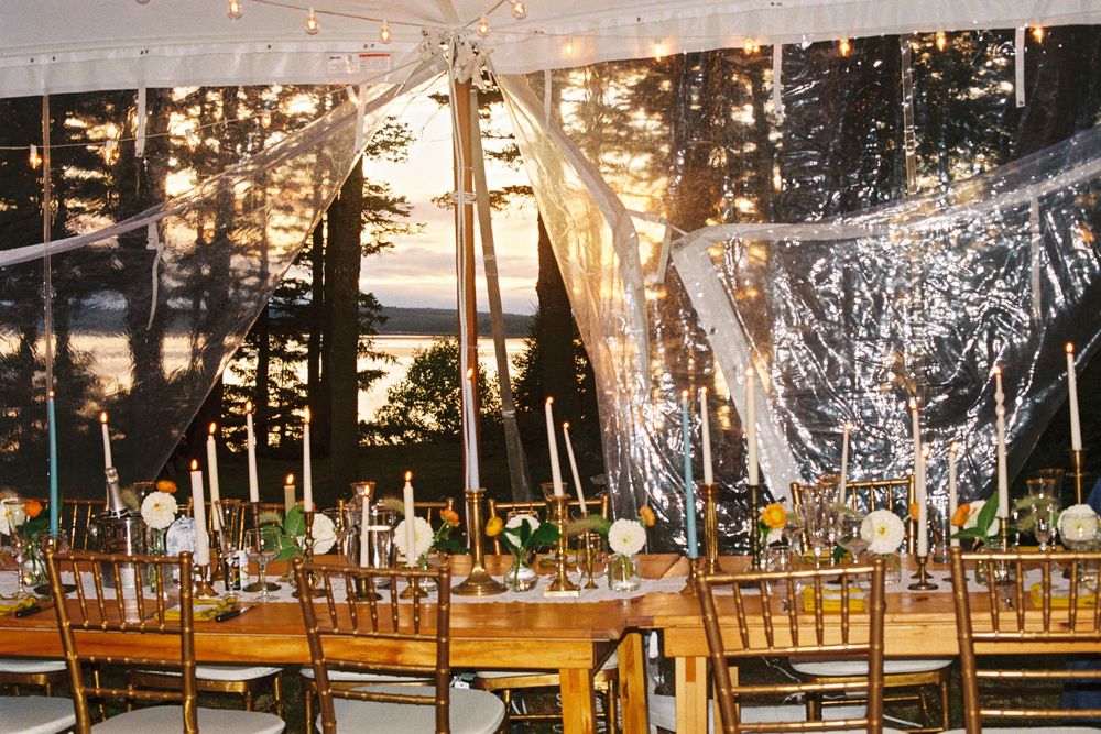 The table settings of a coastal wedding reception with the sun setting through the tent and lighting up the flowers and candles and chairs.