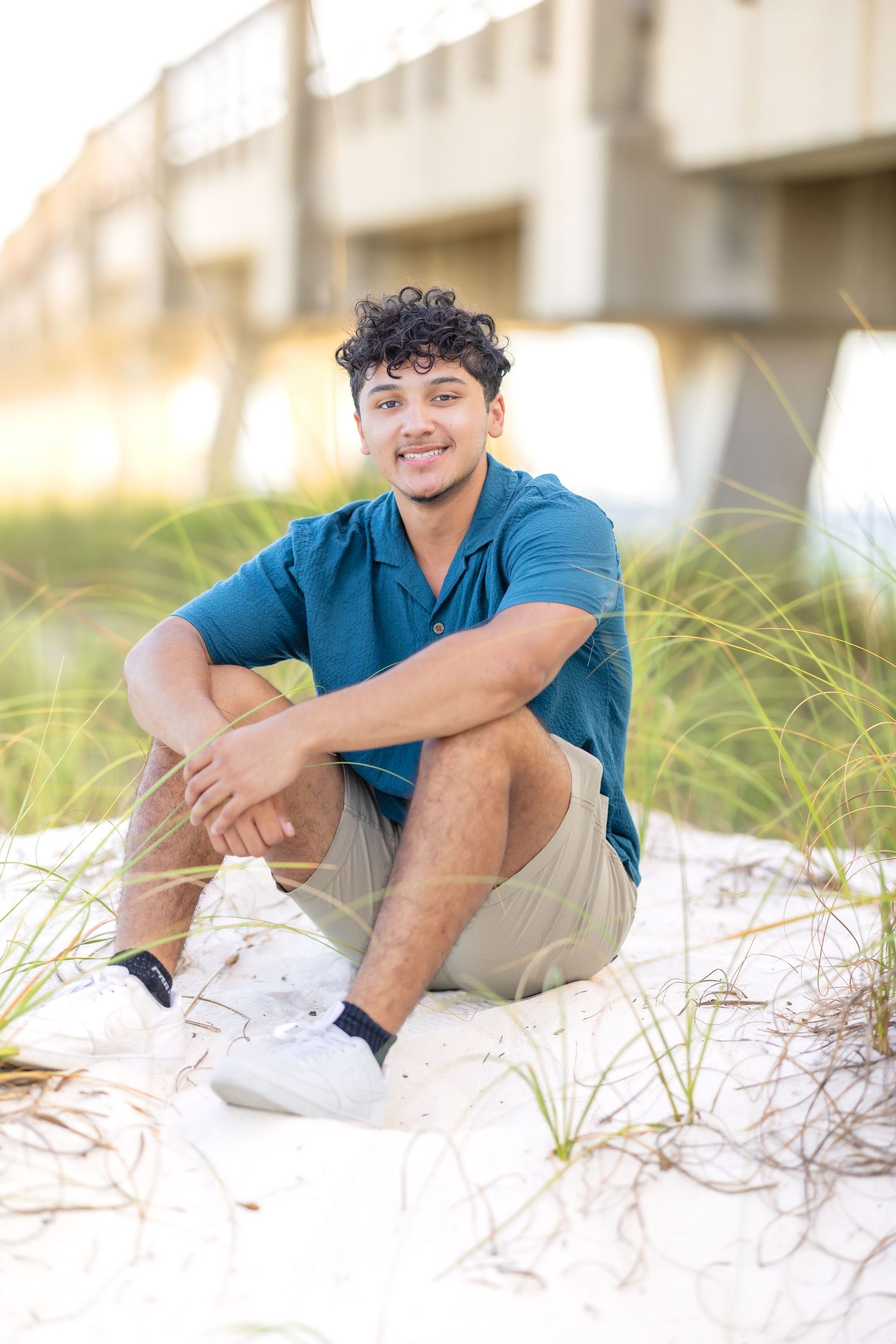 Young person in blue shirt and khaki shorts sits relaxed on sandy beach dunes at sunset.