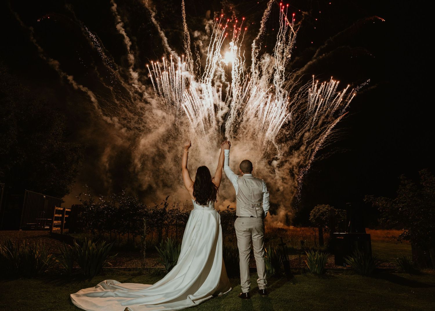 Wedding couple watching spectacular fireworks display during nighttime outdoor celebration with flowing white dress.