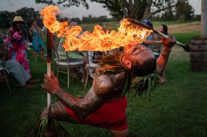 Fire performer breathing flames during an outdoor performance at dusk with chairs and spectators visible in background.