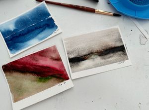 Three abstract watercolor paintings laid out showing blue ocean, black and white landscape, and red mountain scenes.