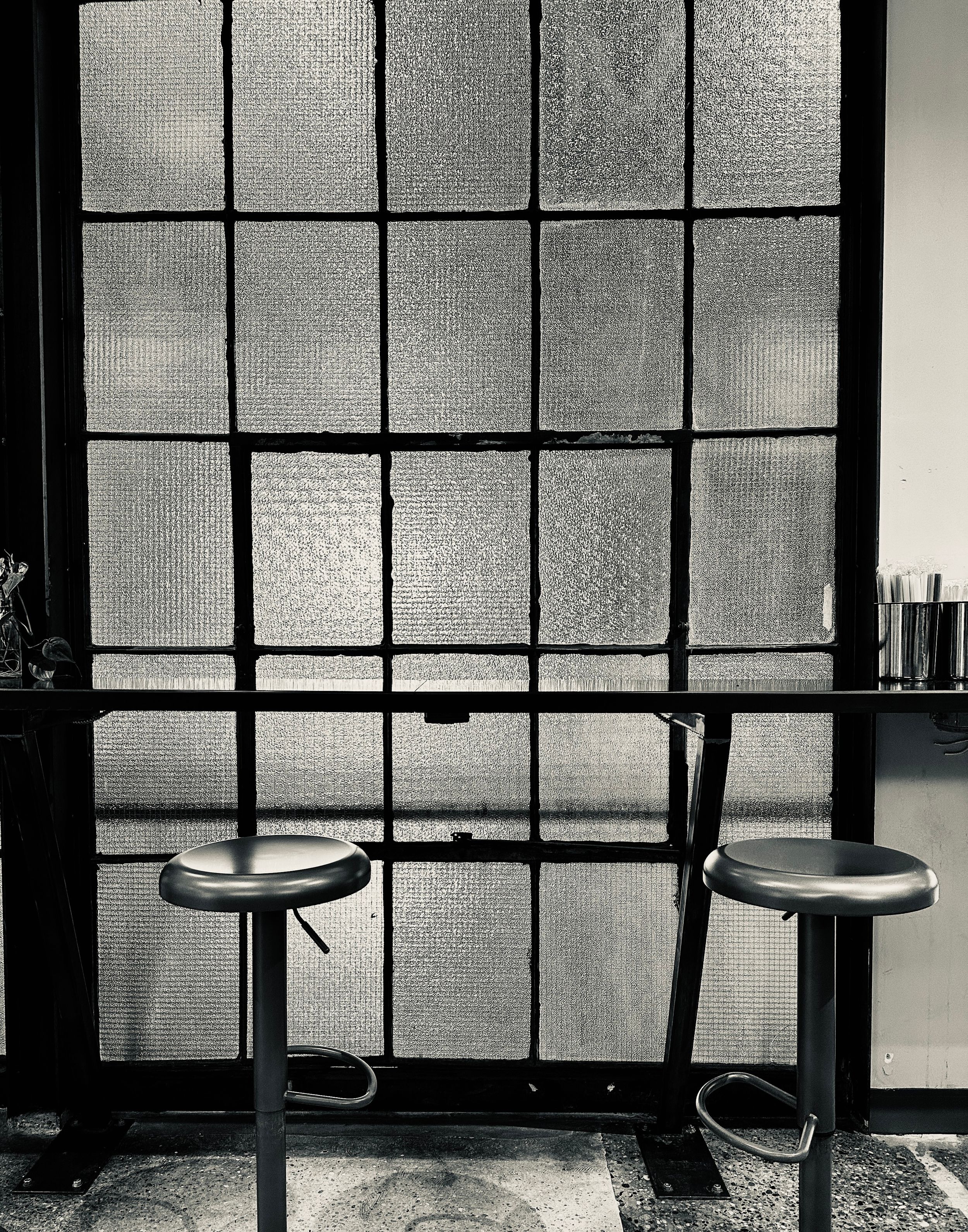 Black and white photo of two bar stools against a geometric window pane with textured glass panels.