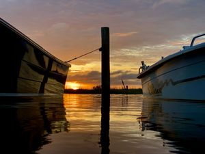 Boats silhouetted against vibrant orange sunset reflecting in calm harbor waters with a wooden post in the foreground.