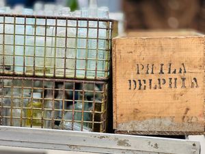 Old wooden shipping box marked 'PHILA DELPHIA' sits next to a metal wire crate containing vintage glass bottles.