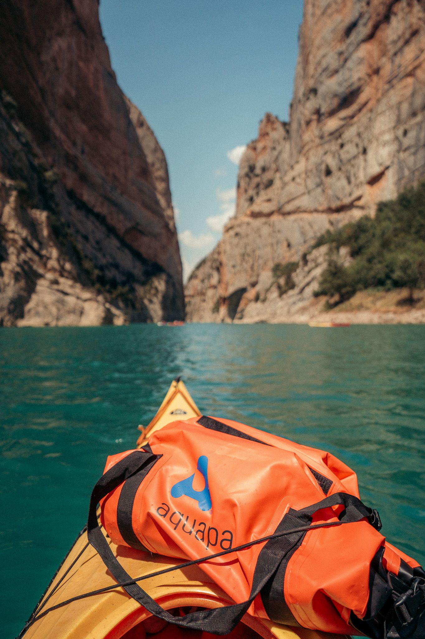 Orange waterproof gear bag secured to kayak in turquoise waters between canyon walls.