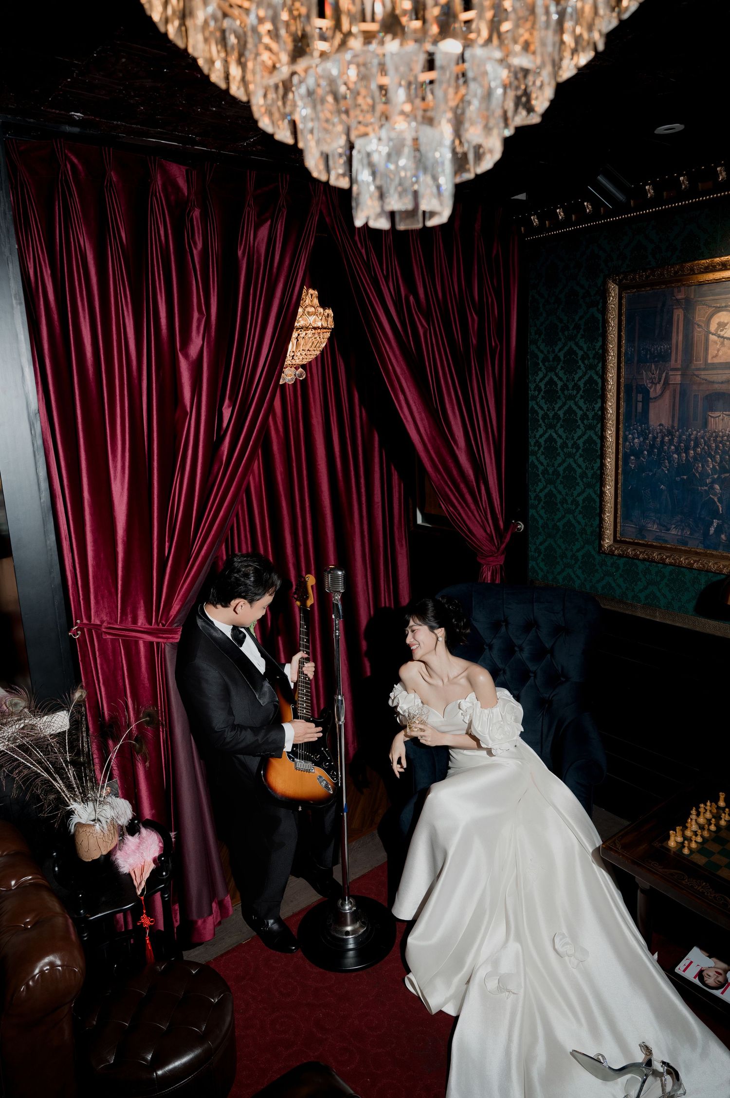 Bride in white gown and groom in tuxedo share a moment under a crystal chandelier with red velvet curtains.
