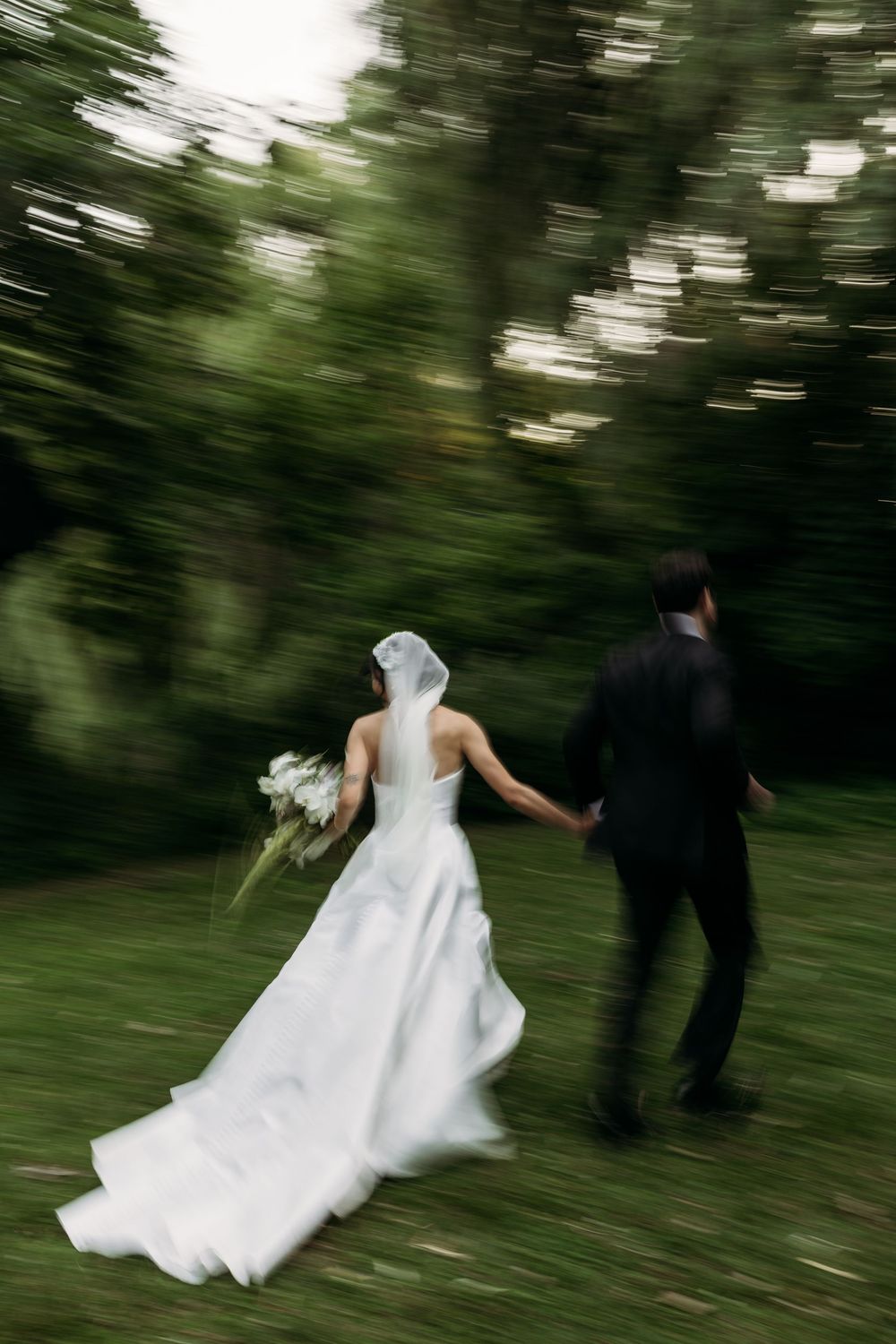 Motion-blurred photo of newlyweds running through a green garden, with the bride's white dress flowing behind them.