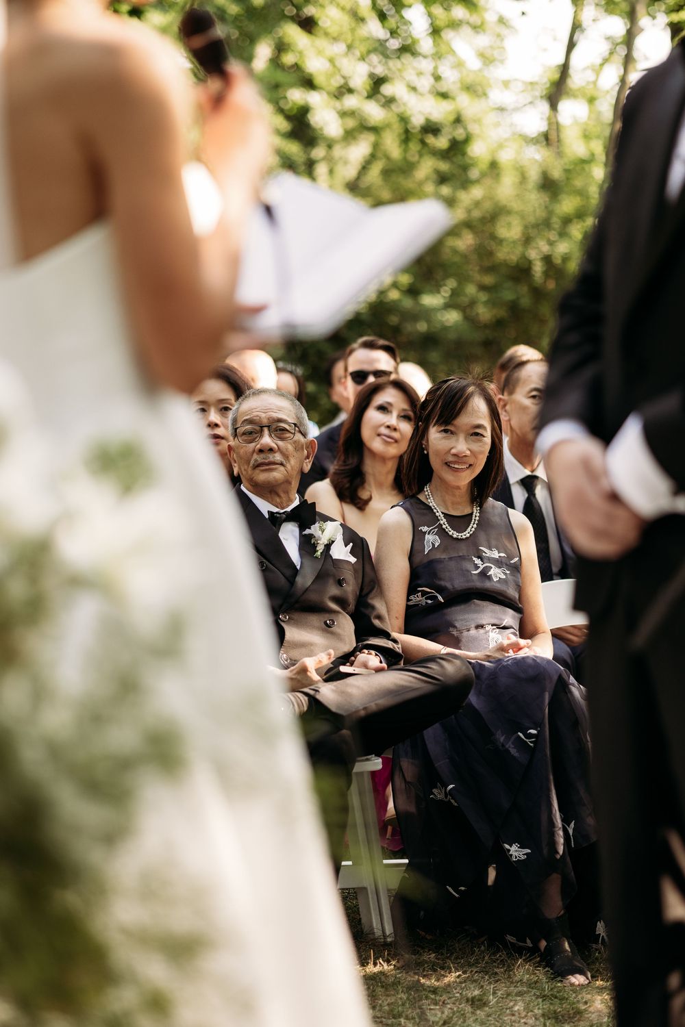 Wedding guests in formal attire seated outdoors watching the ceremony through soft focus trees and greenery.