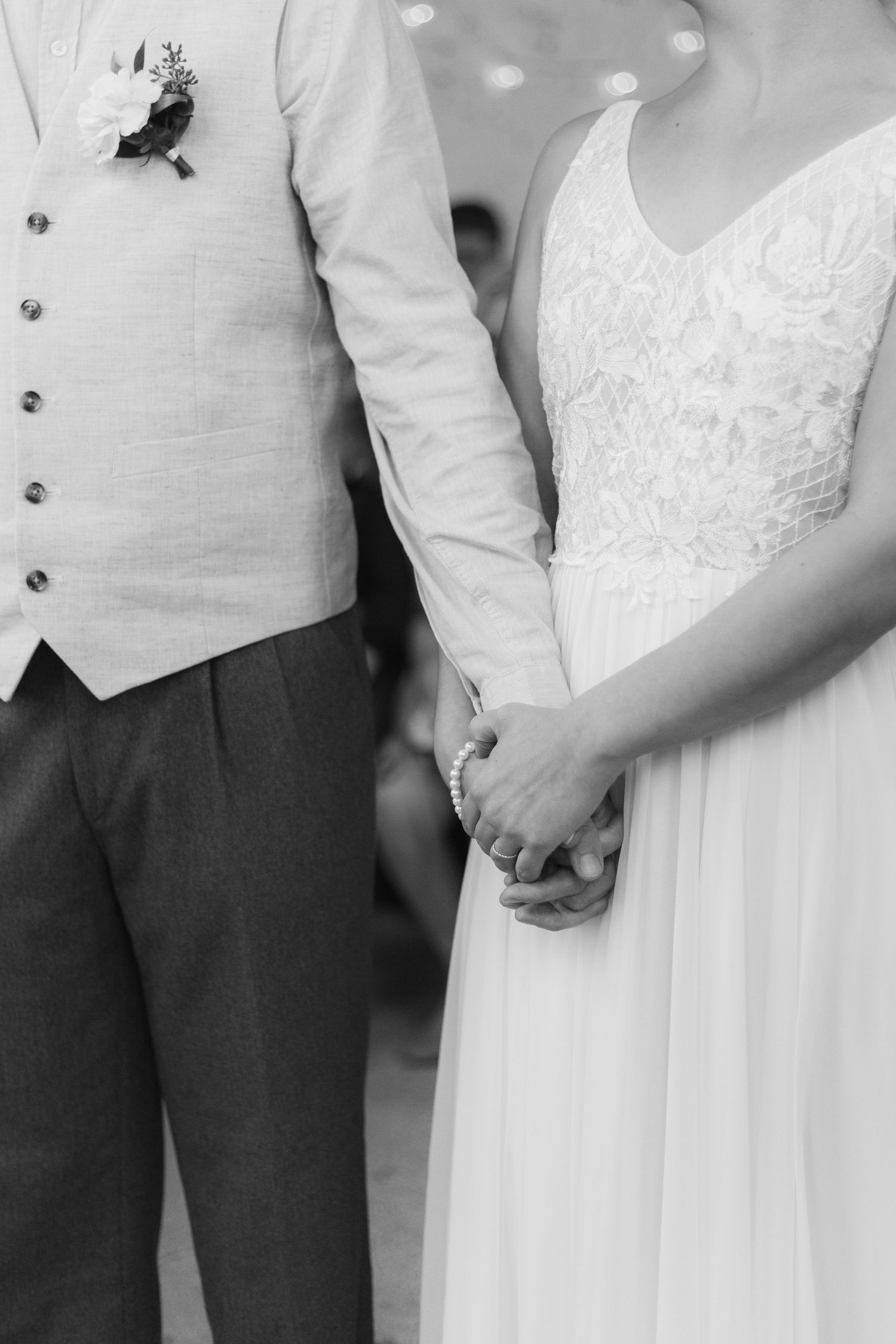 Black and white photograph of newlyweds holding hands, showing bride in white lace dress and groom in grey vest.